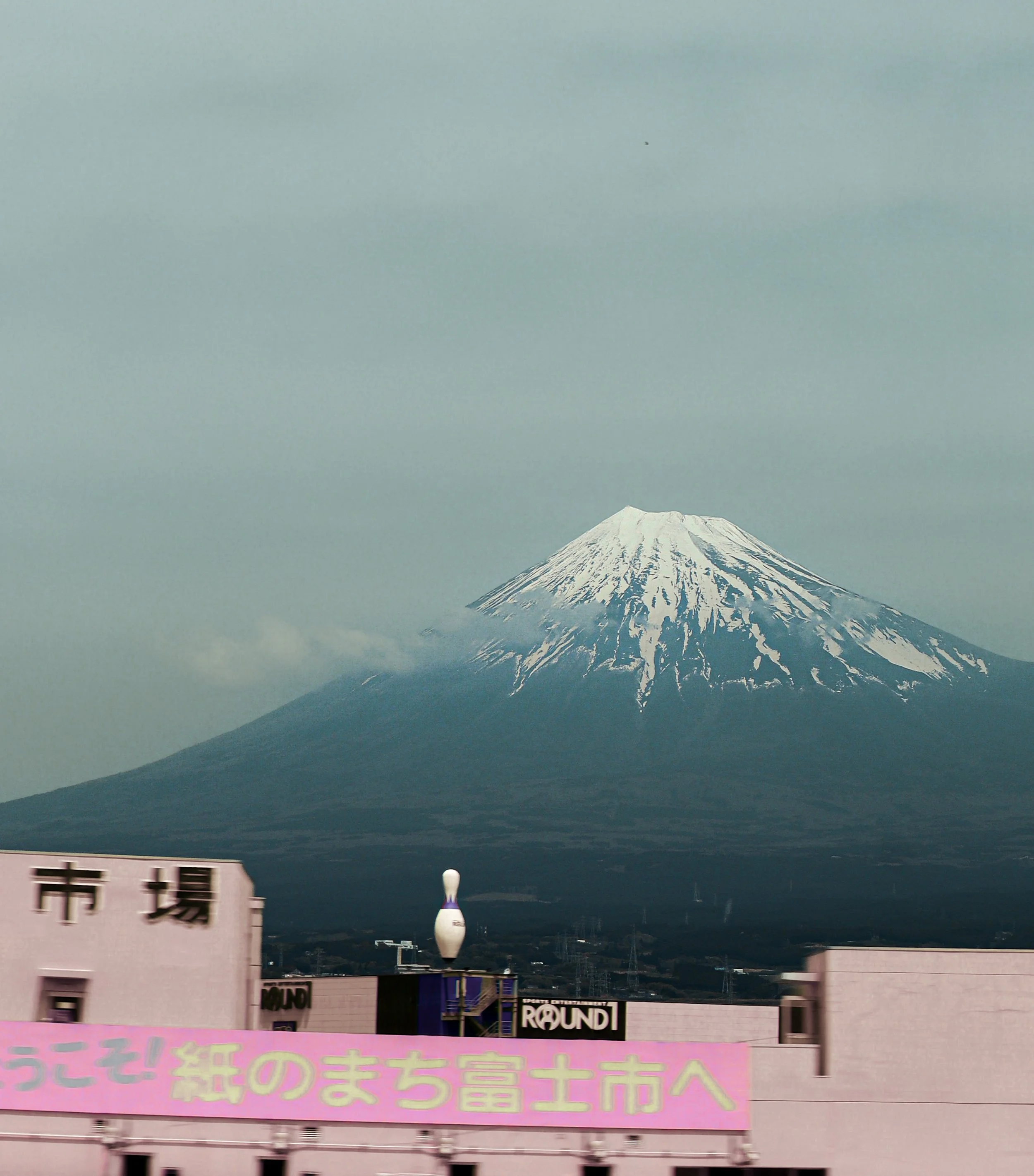 A mountain with snow on top, likely Mount Fuji, seen through an urban area with buildings and signs, including a pink sign with Japanese characters in the foreground.