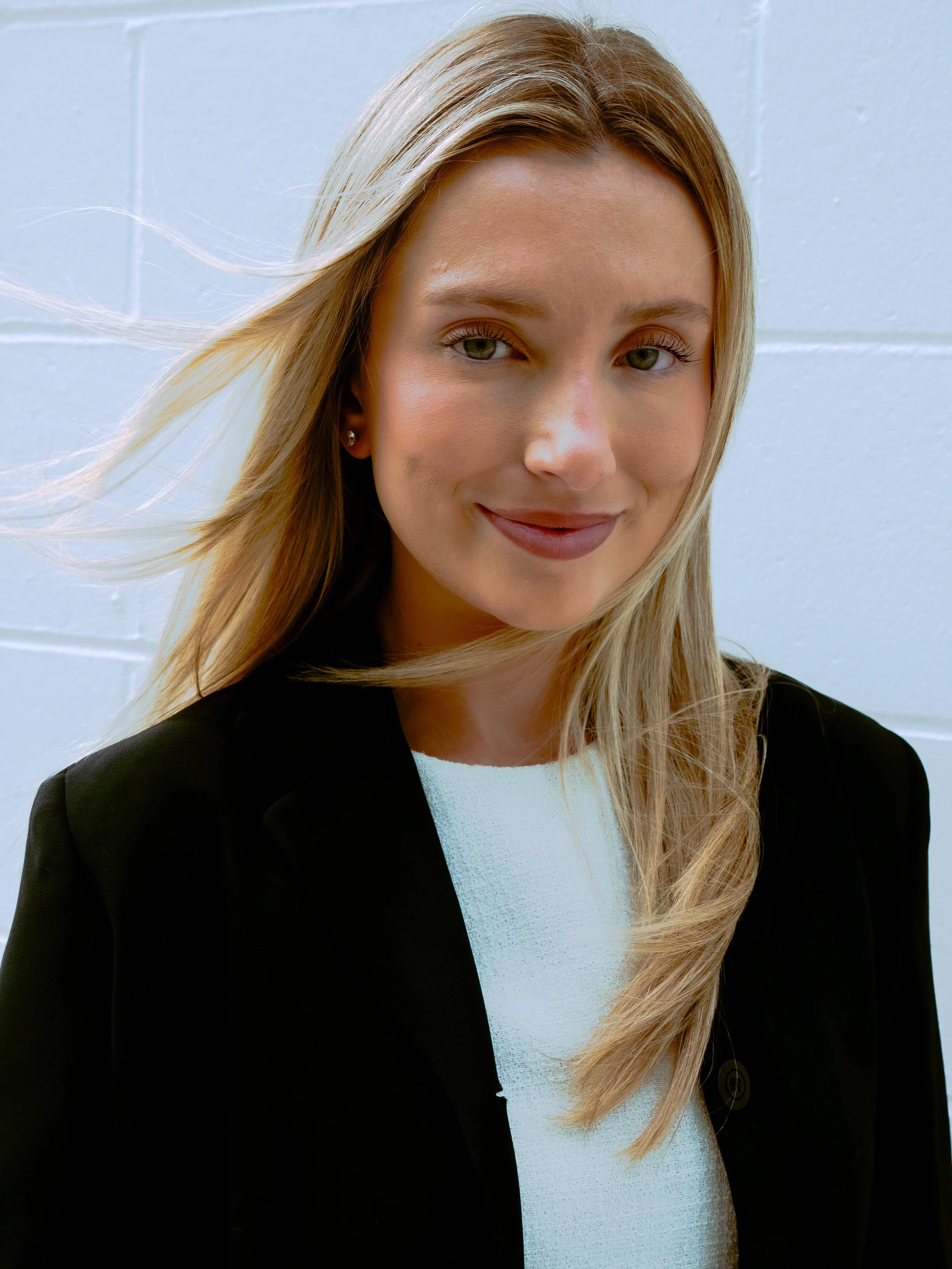 Close-up of a smiling woman with blonde hair wearing a black blazer, white top, and small earrings, standing against a light blue wall.