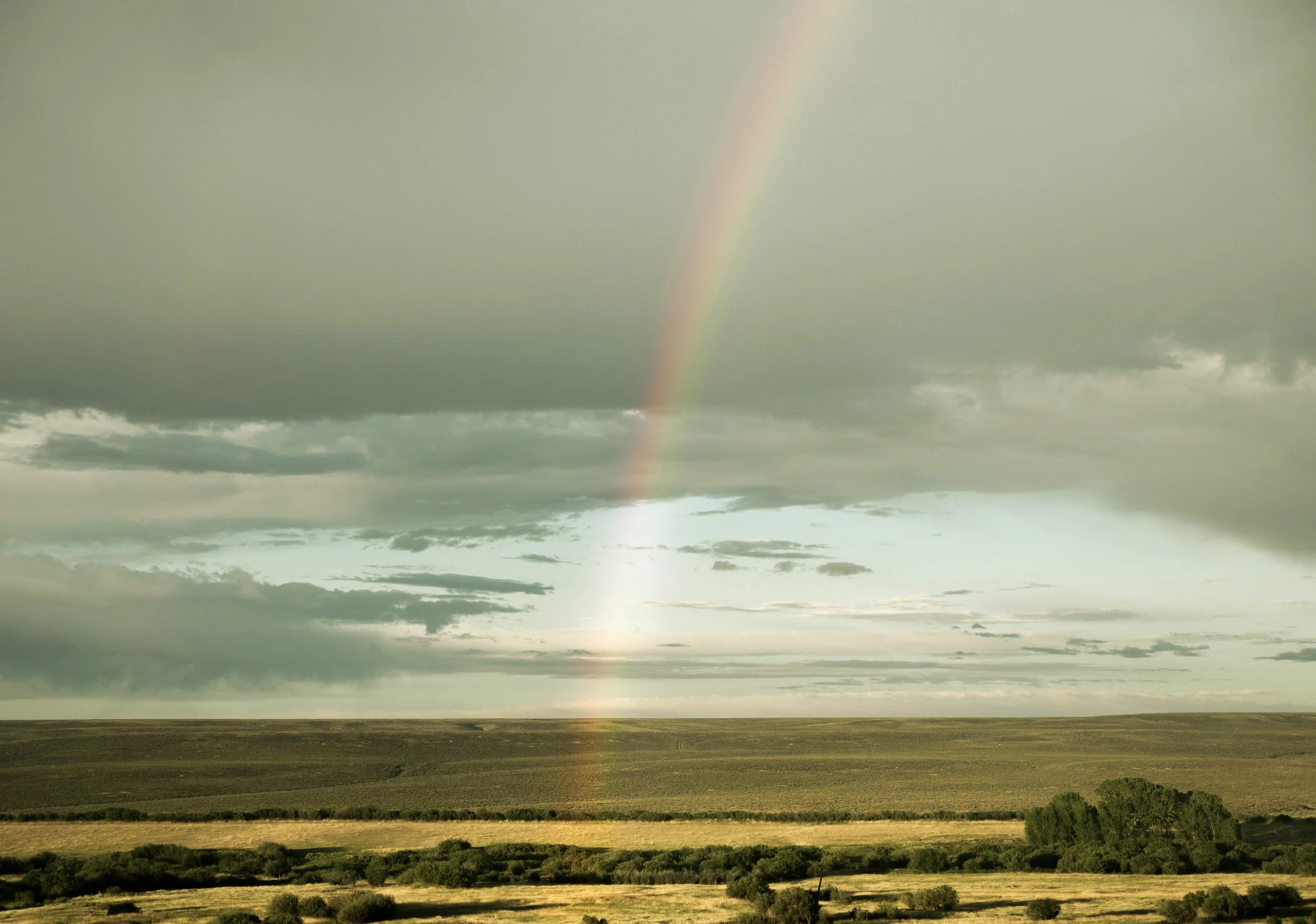 A landscape view of open fields under a cloudy sky with a faint rainbow visible in the distance.