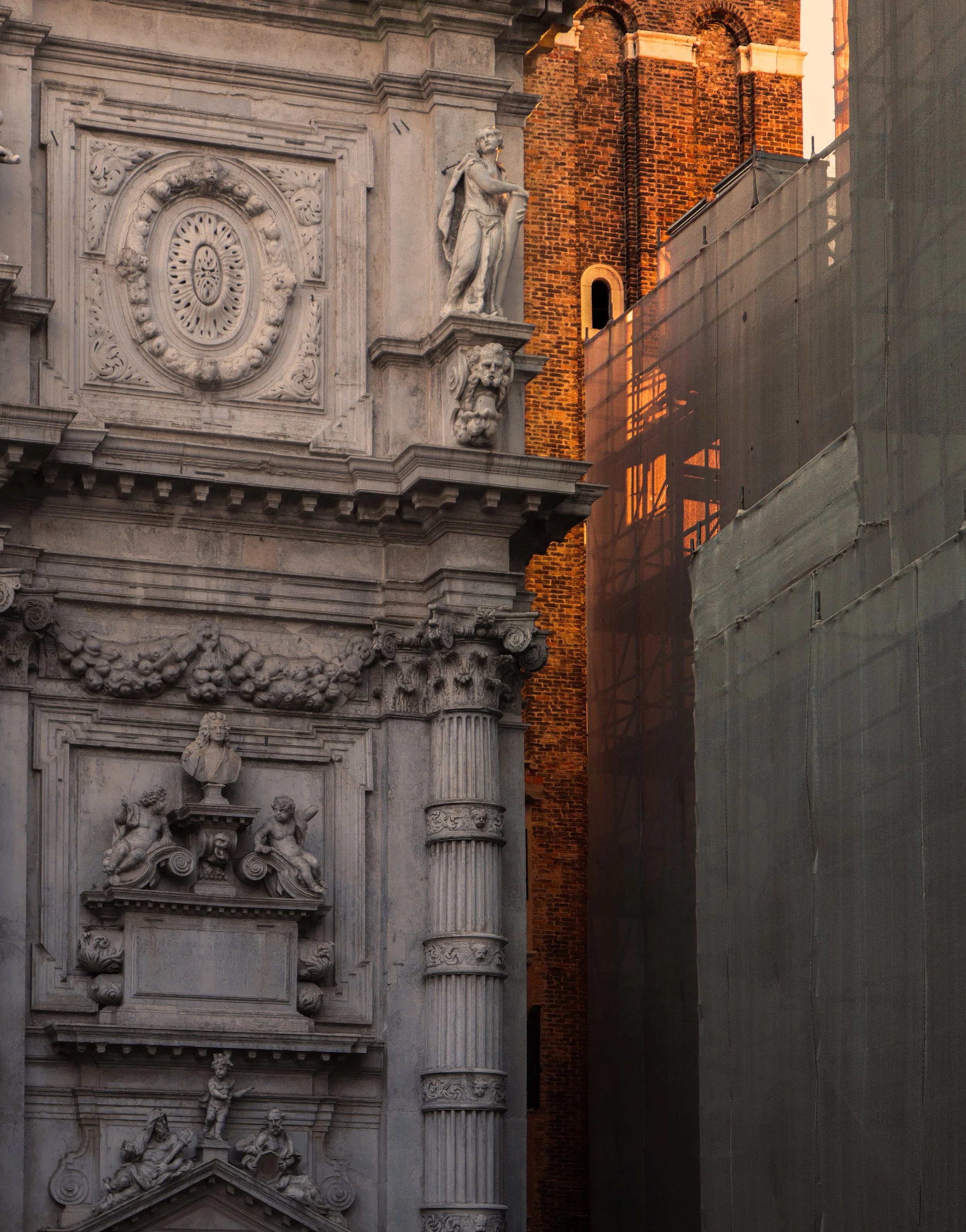 Close-up view of ornate stone facade with sculptures, columns, and decorative details, beside a brick building and a construction site with scaffolding covered in protective green mesh at sunset.