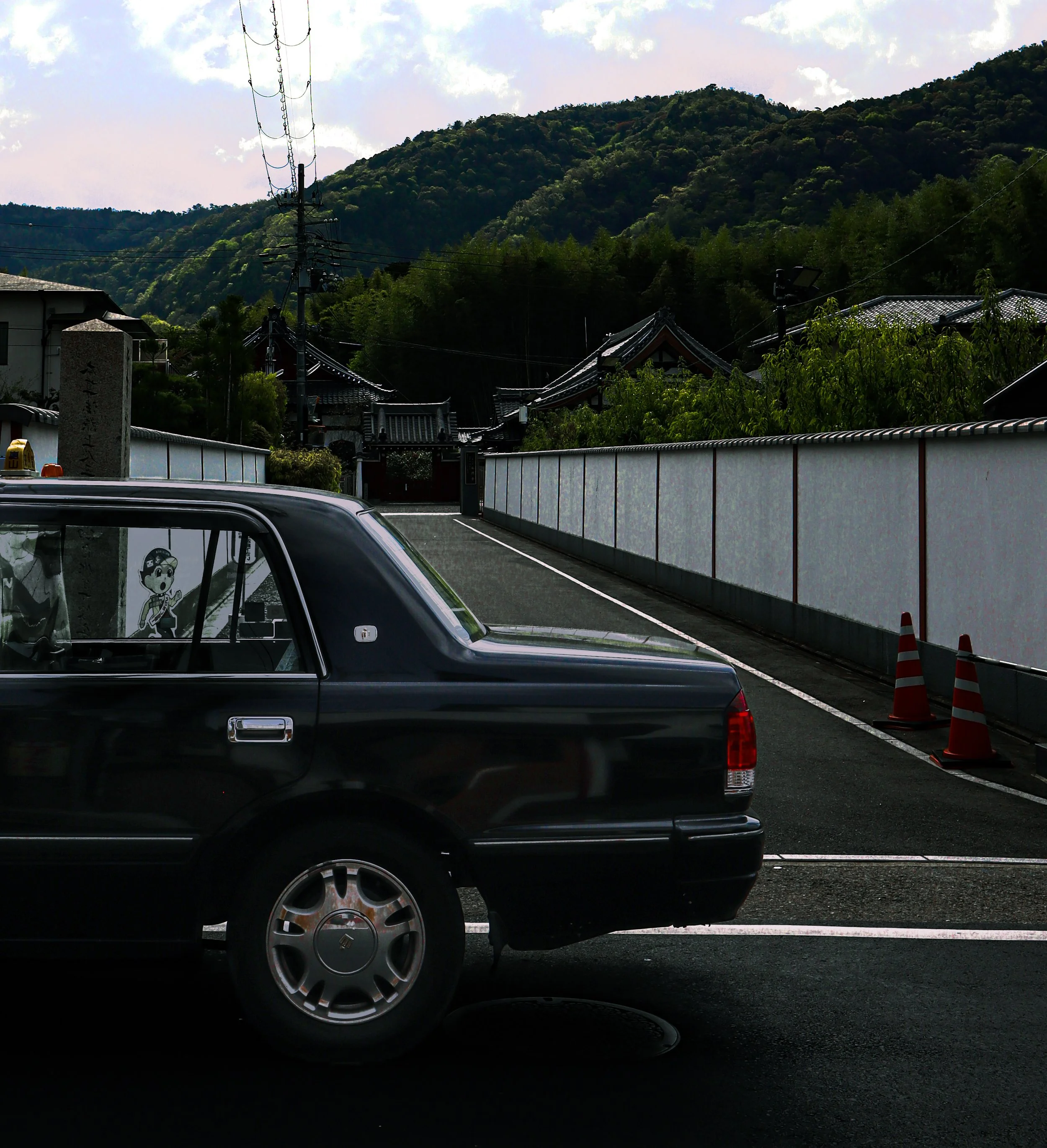A black car parked in front of a white wall with Japanese architecture rooftops visible behind it. There are orange and white traffic cones on the street beside the car, and mountains with lush green trees in the background.
