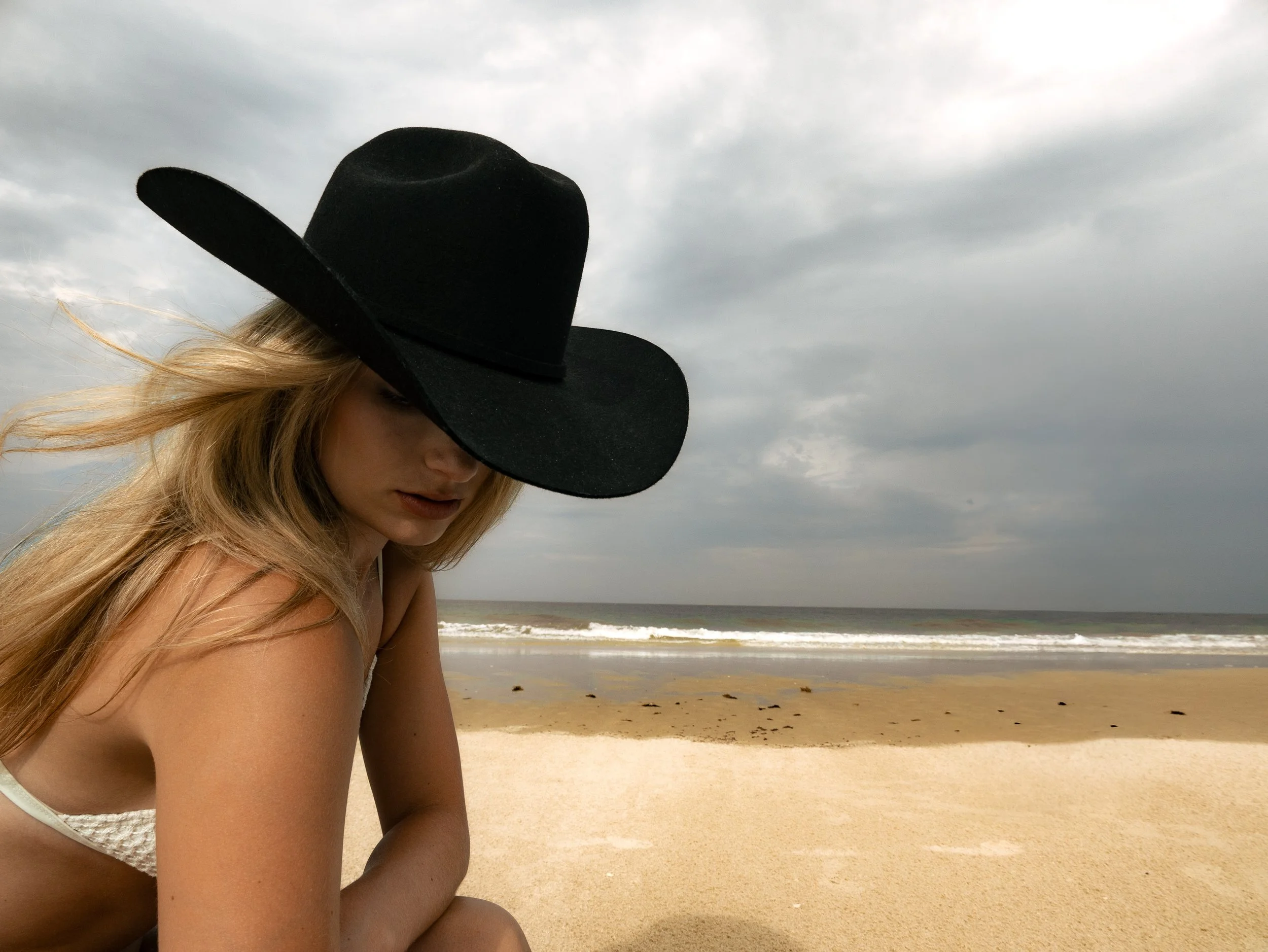 A woman with long blonde hair wearing a large black hat and white clothing, sitting on a sandy beach with the ocean and cloudy sky in the background.