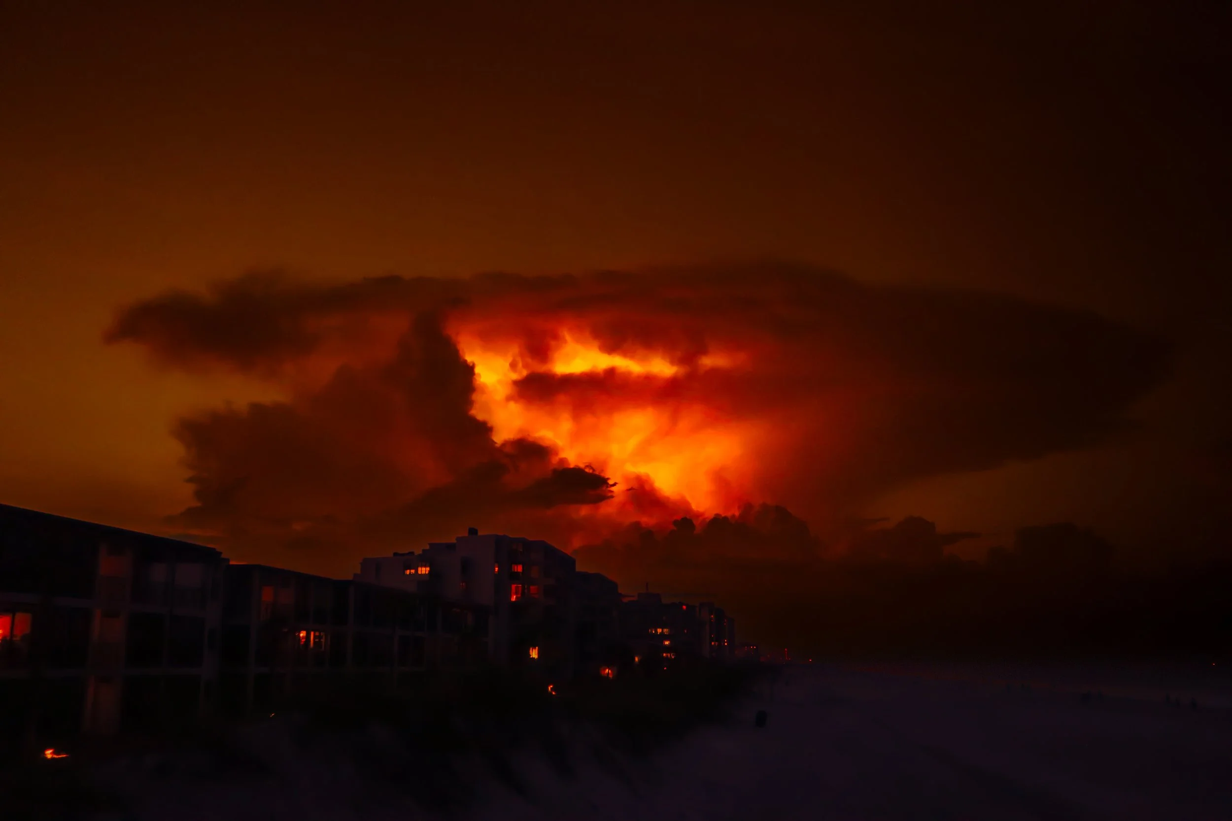 A dramatic sunset with fiery orange and red clouds over buildings and a beach.