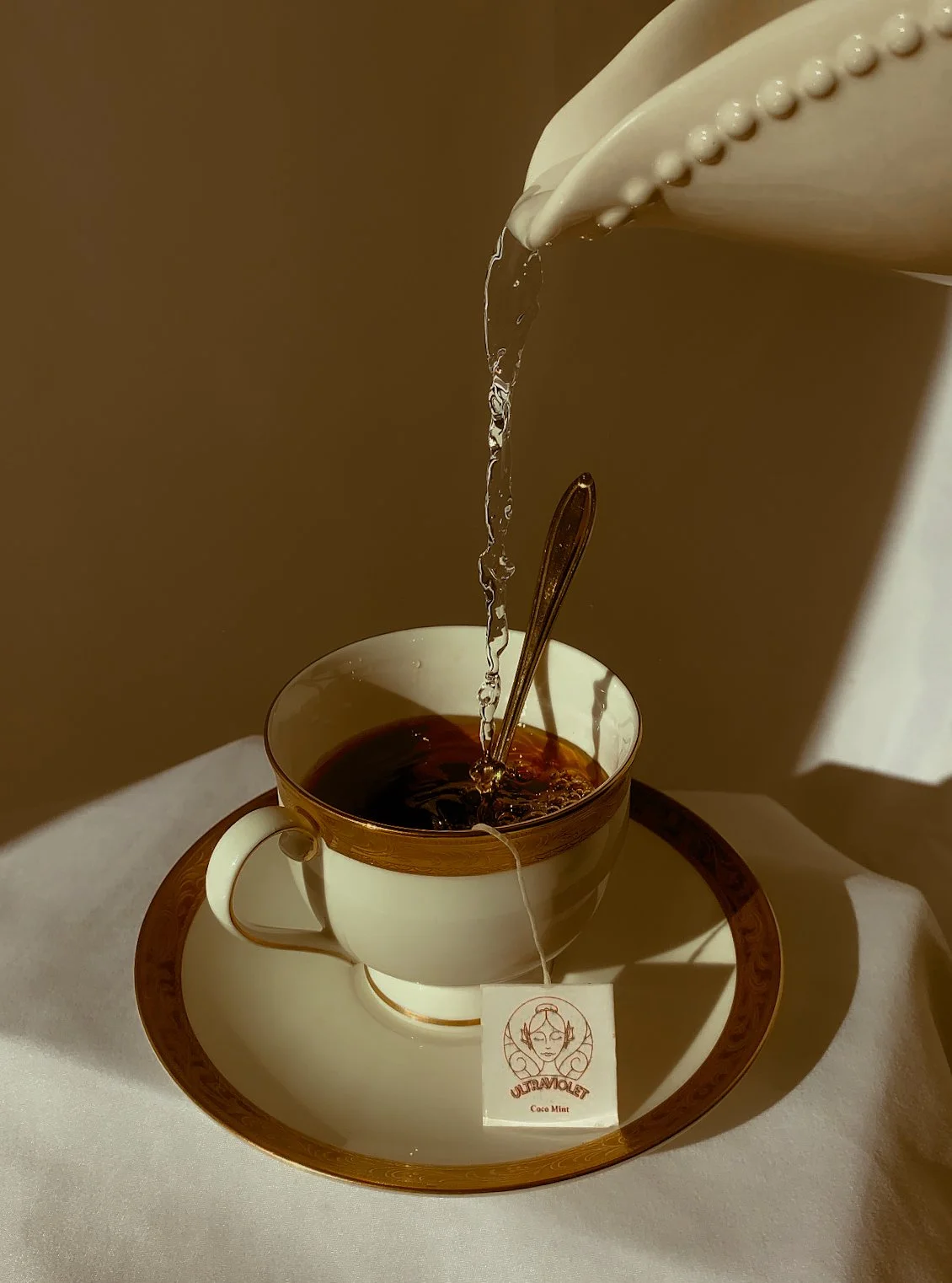 A white teacup with gold trim placed on a matching saucer and tray, with a tea bag submerged in hot tea. A spoon rests inside the cup, and water is being poured from a white teapot into the cup. The tea bag label reads "ULTRAVIOLET Coco Mint." The sc