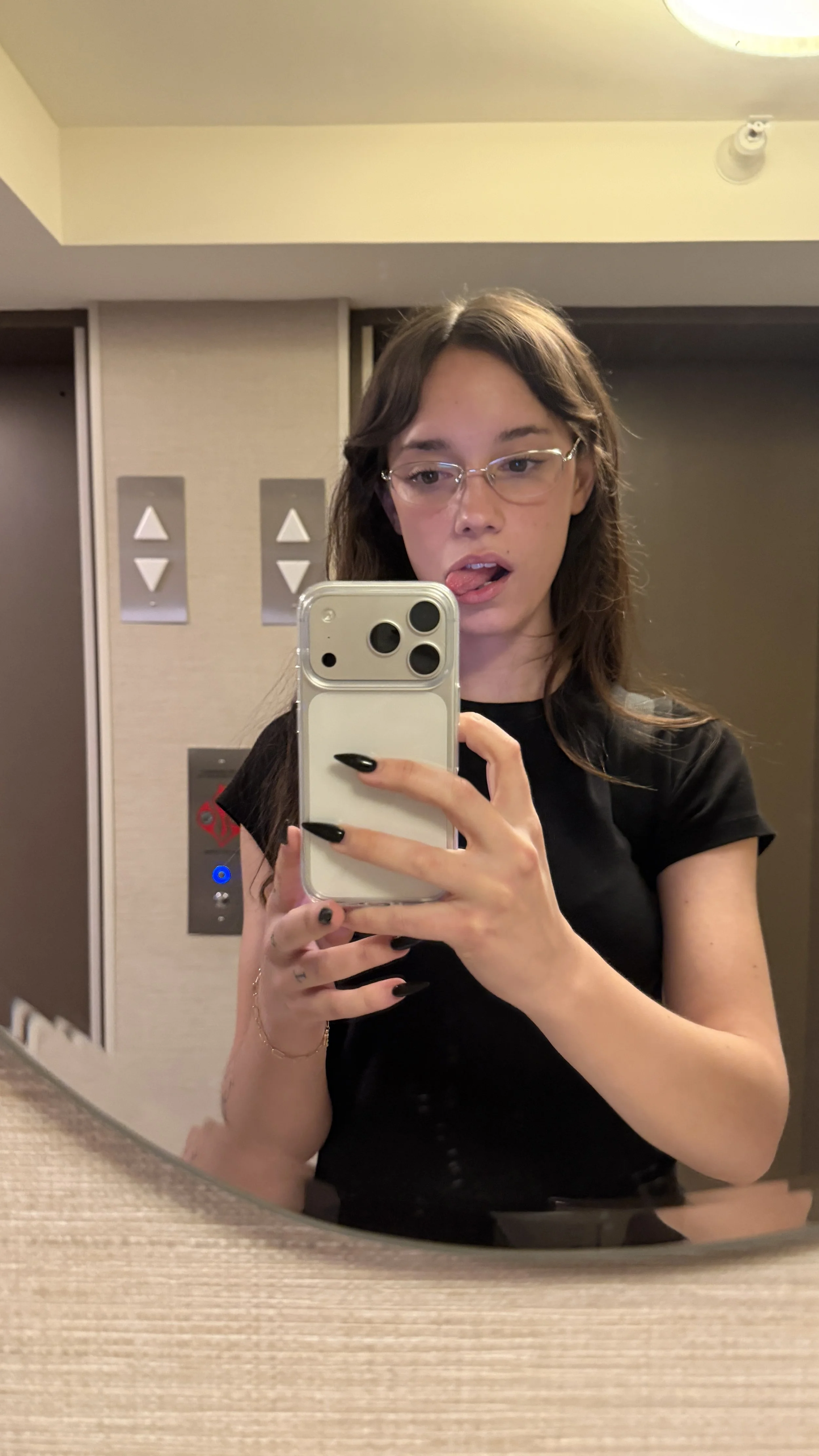 A young woman with glasses and long brown hair taking a mirror selfie in an elevator, sticking her tongue out slightly, wearing a black top, with the elevator buttons and a ceiling light visible in the background.