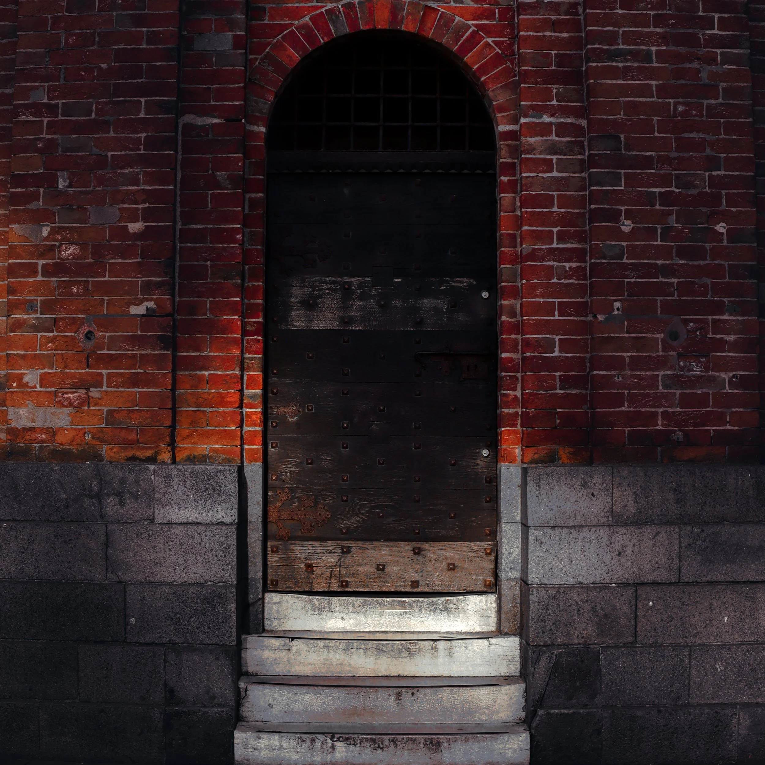 A brick building with a black wooden door that has a rounded arch at the top, featuring a metal grid covering a small dark window. The bricks are red and weathered, with some mortar and paint peeling.
