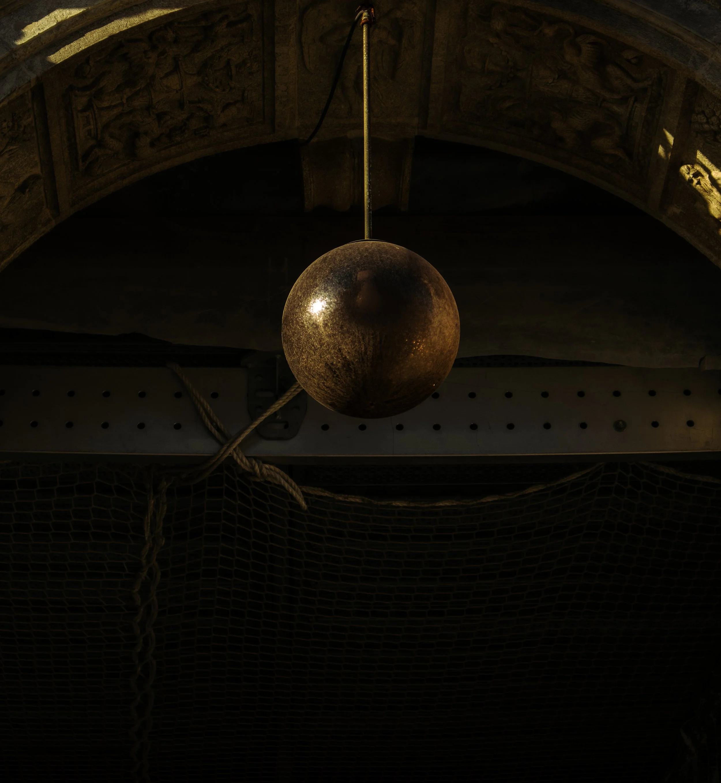 A spherical object hangs from the ceiling, with a textured, metallic surface, suspended by a yellow cord. The ceiling above features ornate carved stone artwork, possibly depicting mythical or historical scenes. The background includes netting and ot