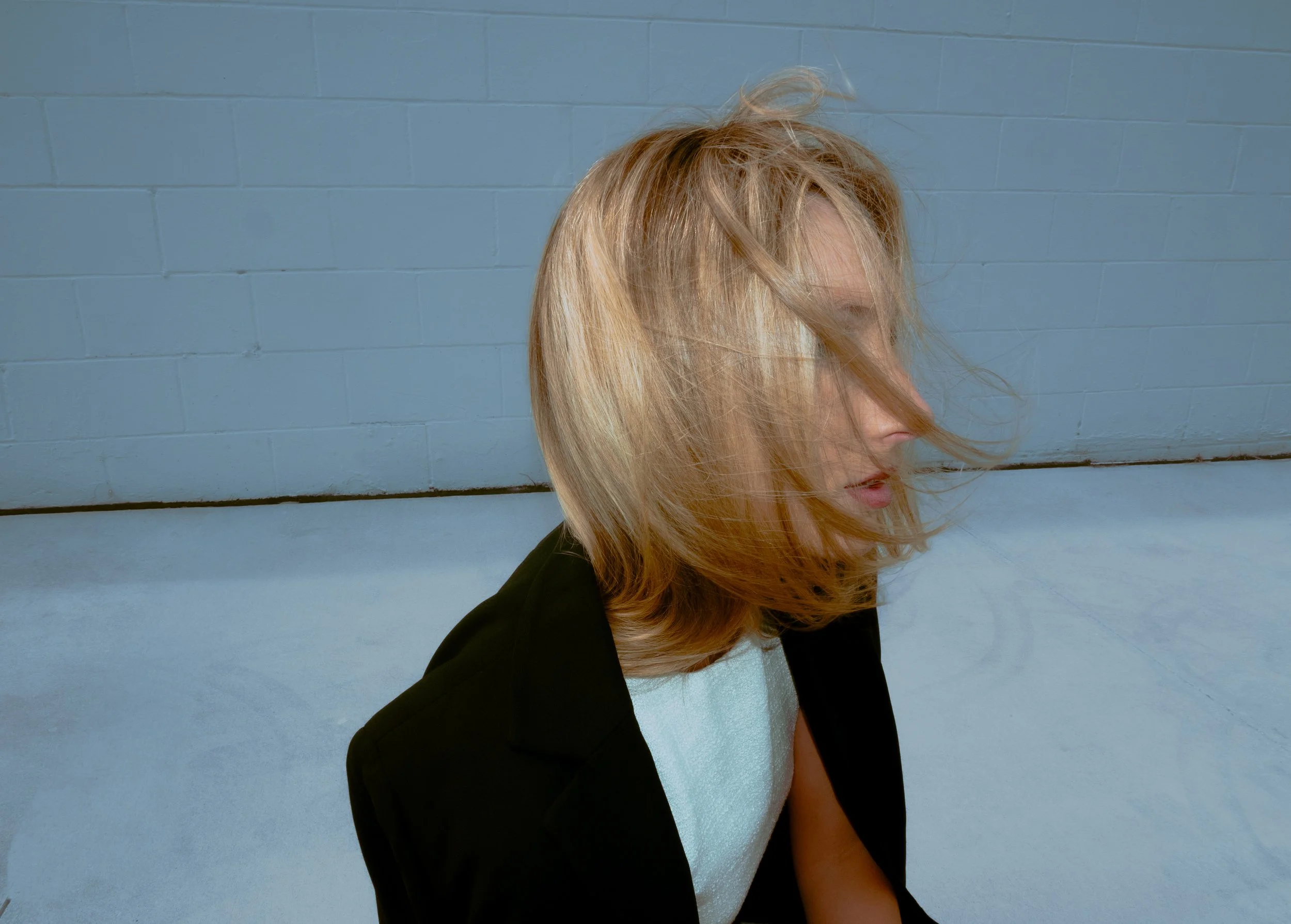 A woman with blonde hair turning to the side, standing against a blue painted brick wall.