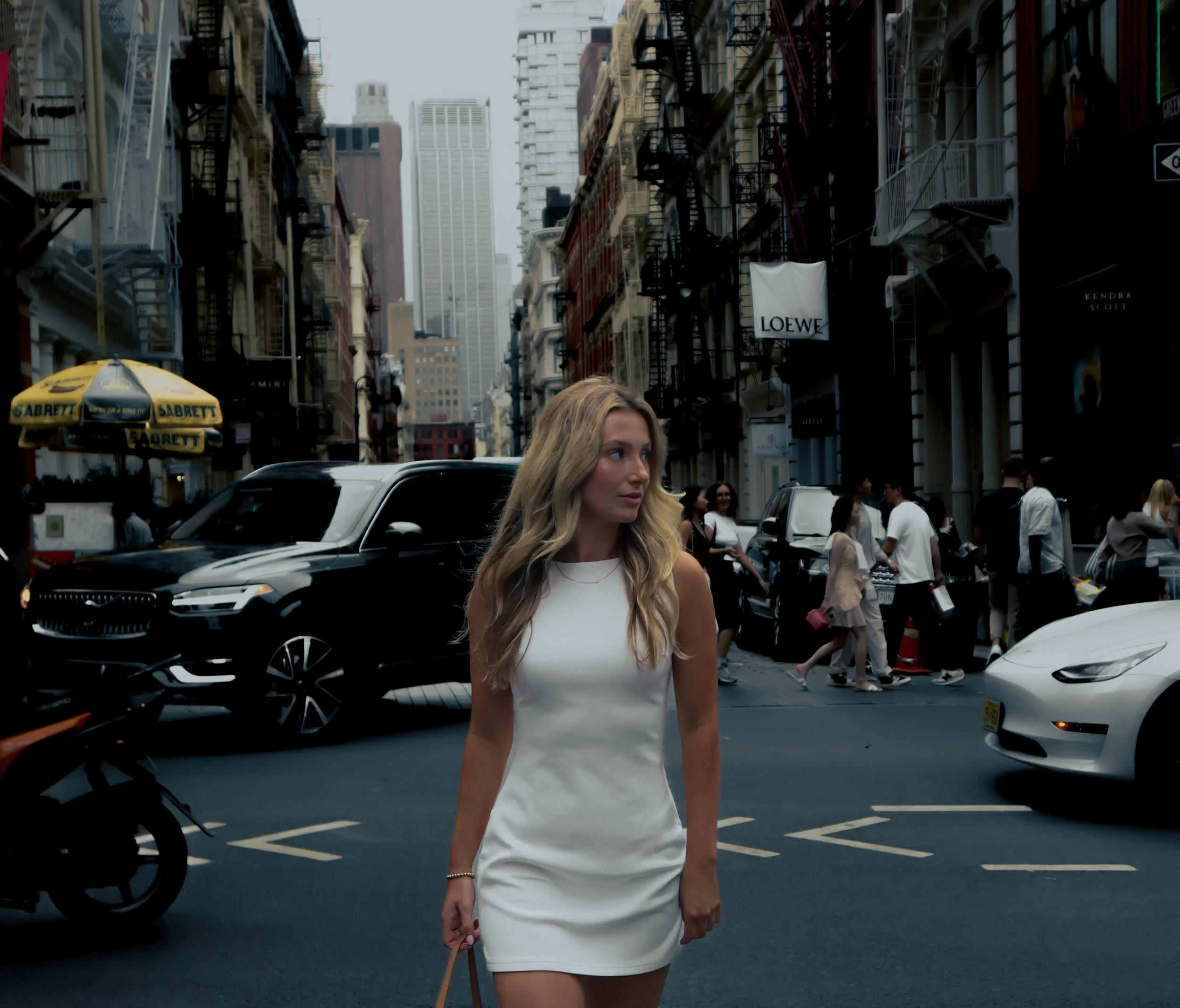 A woman with blonde hair wearing a white sleeveless dress walking on a busy city street with cars and people. There are tall buildings and fire escapes visible on the sides of the street. A yellow umbrella with the Sabrett logo is in the foreground.