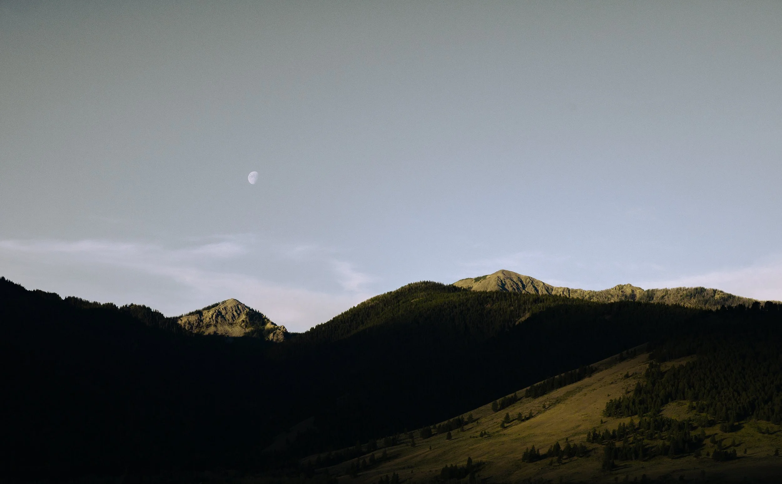 Mountain landscape with dark forested hills in the foreground, lighter hills in the middle ground, and a clear sky with a visible moon in the background.