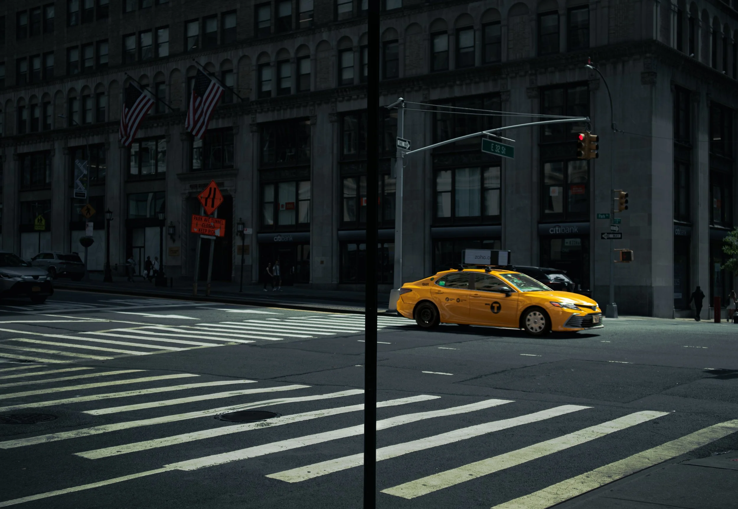 A city street scene at the intersection of E 32nd Street with a yellow taxi cab stopped at a red traffic light. There are multiple American flags hanging from a building in the background, which has a sign for Citibank and several pedestrians walking