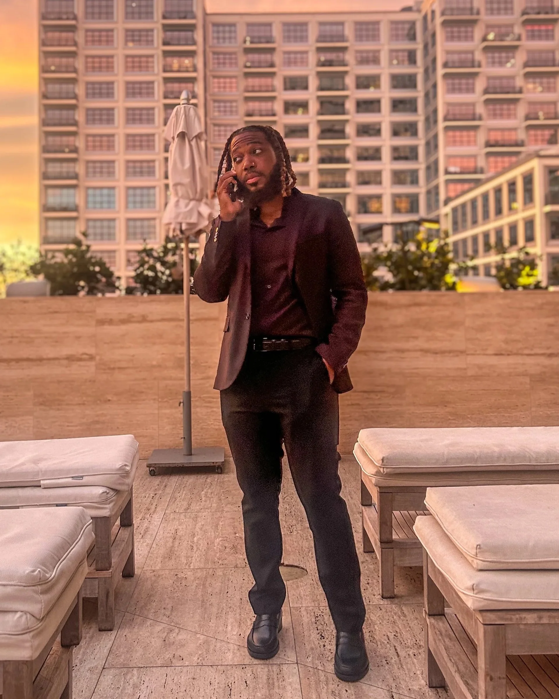 A man with dreadlocks wearing a black blazer and black pants stands on a terrace at sunset, talking on his phone. The terrace has mattresses on wooden frames and an open parasol, with a cityscape of tall buildings in the background.