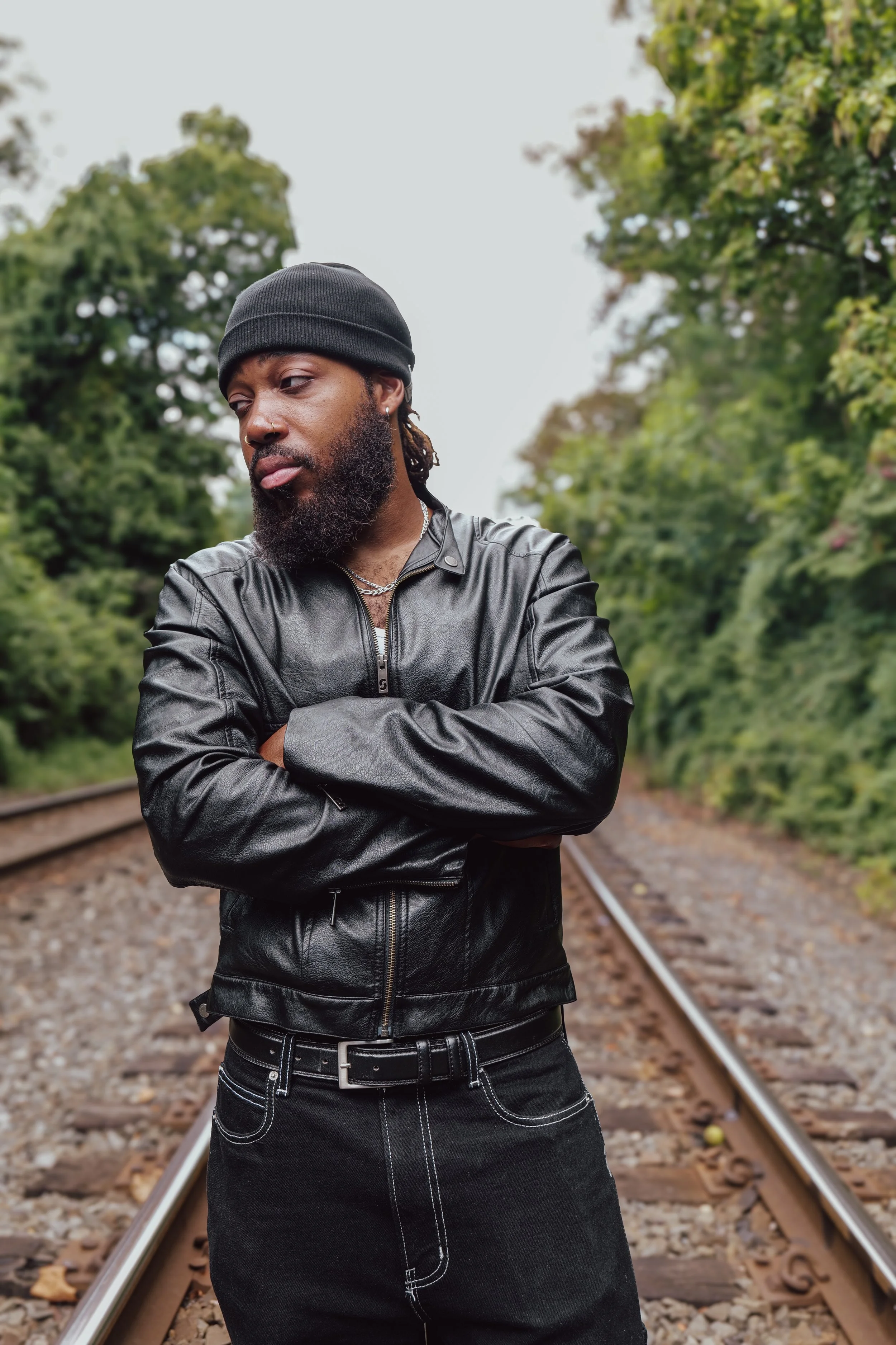 A man wearing a black beanie, a black leather jacket, and black jeans stands with crossed arms on a railroad track surrounded by green trees.
