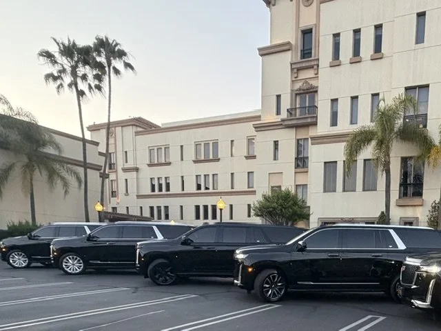Black luxury SUVs parked in a parking lot outside a multi-story beige building with palm trees.