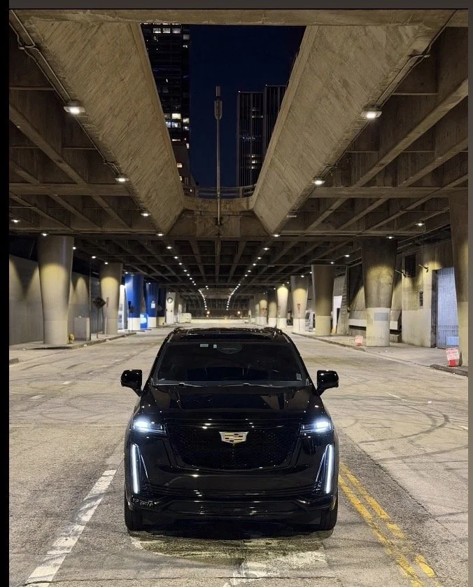 Black Cadillac SUV parked under a highway bridge at night in an urban area.