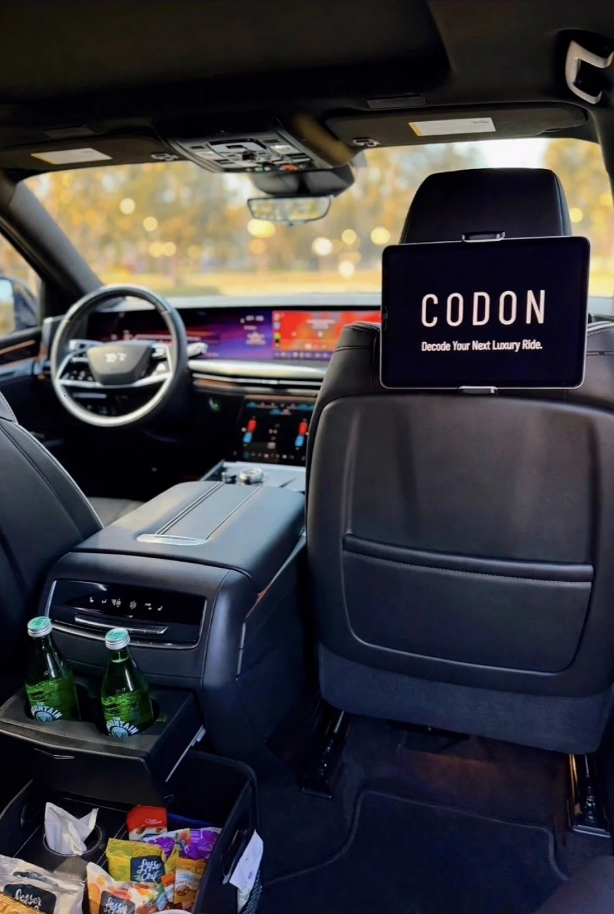 Interior of a luxury vehicle with a digital dashboard, a screen on the back of the front seat displaying 'CODON' and 'Decode Your Next Luxury Ride,' bottles of water in the center console, and snacks in a tray below.