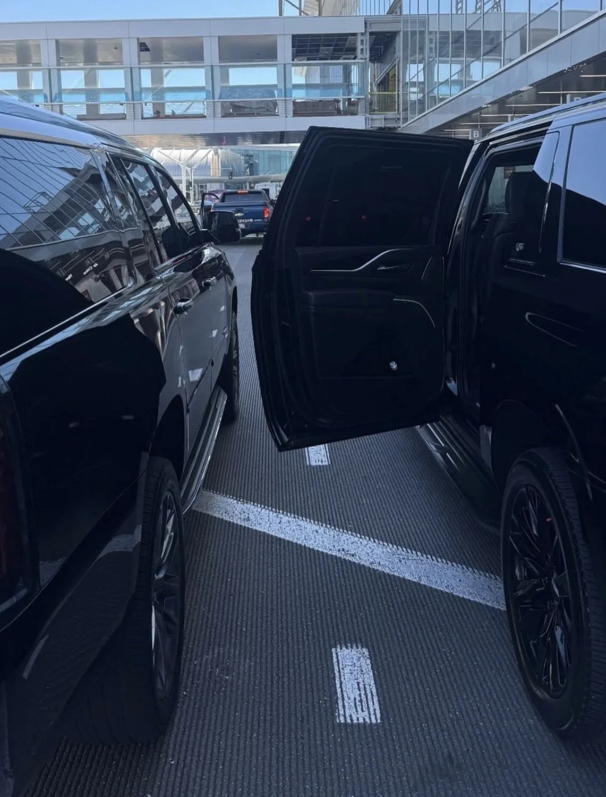 Multiple black SUVs parked at an airport terminal, with one vehicle's door open, and a glass building in the background.