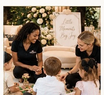 Two women sitting on the floor with young children, engaging in an activity, with a floral backdrop and a sign that says 'Little Moments Big Joy' in the background.