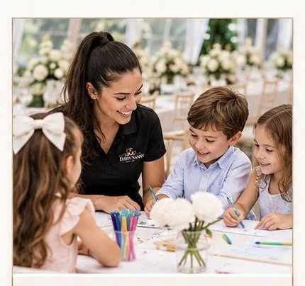 A woman with dark hair in a ponytail smiling while engaging with three children at a table decorated with flowers, markers, and paper, in a bright indoor setting.