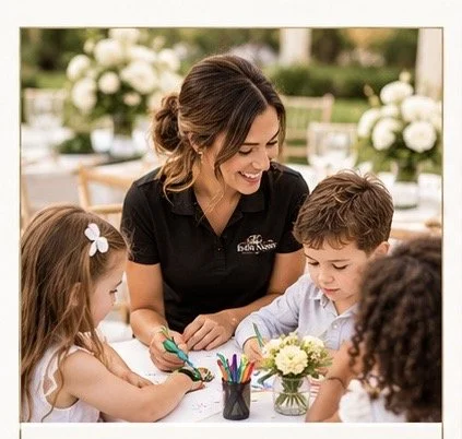A woman and three children sitting at a table with arts and crafts supplies, outdoors with flowers in the background.