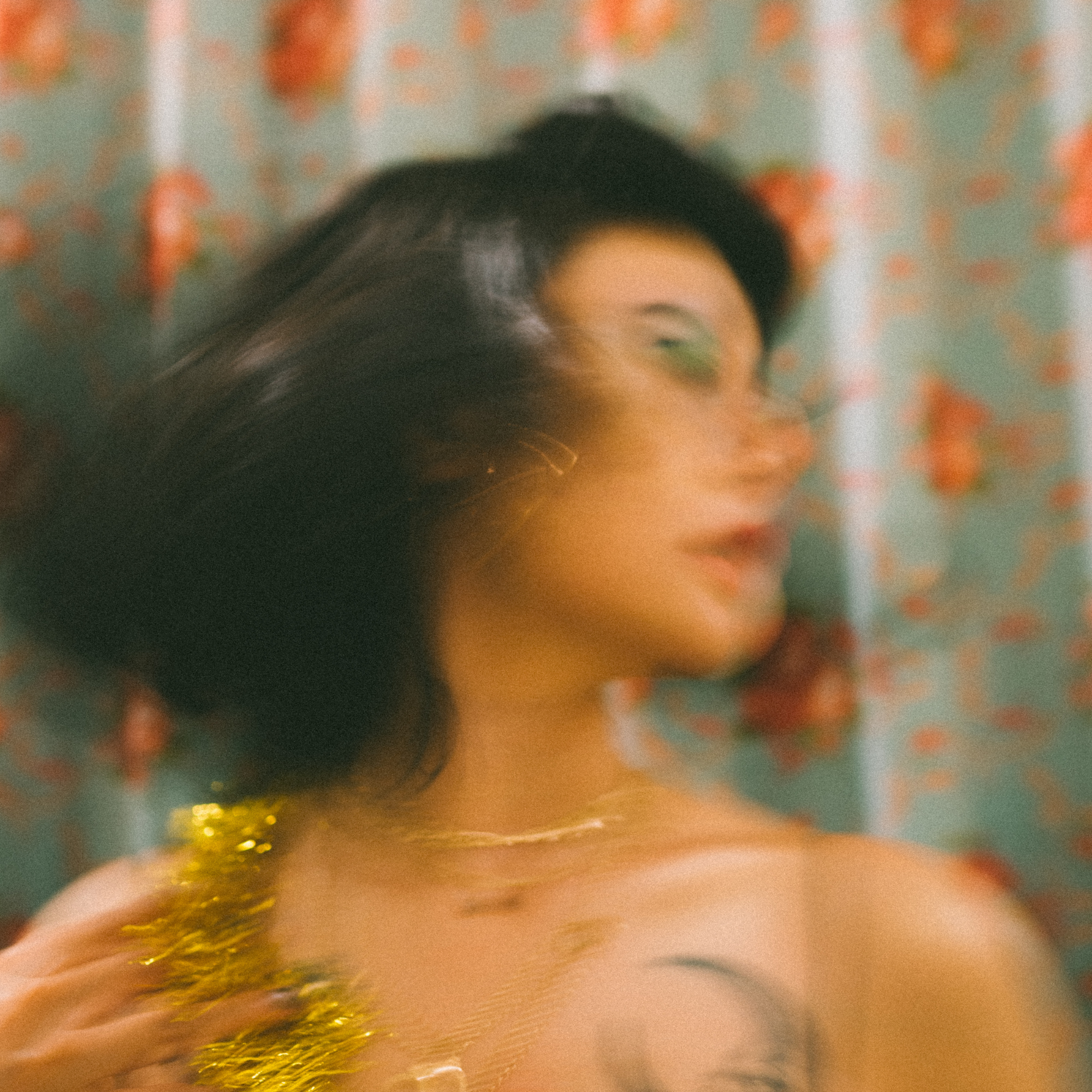 A close-up of a person's head with black hair, green eye makeup, and a gold tinsel bracelet on their wrist, set against a background with orange and green polka dots.