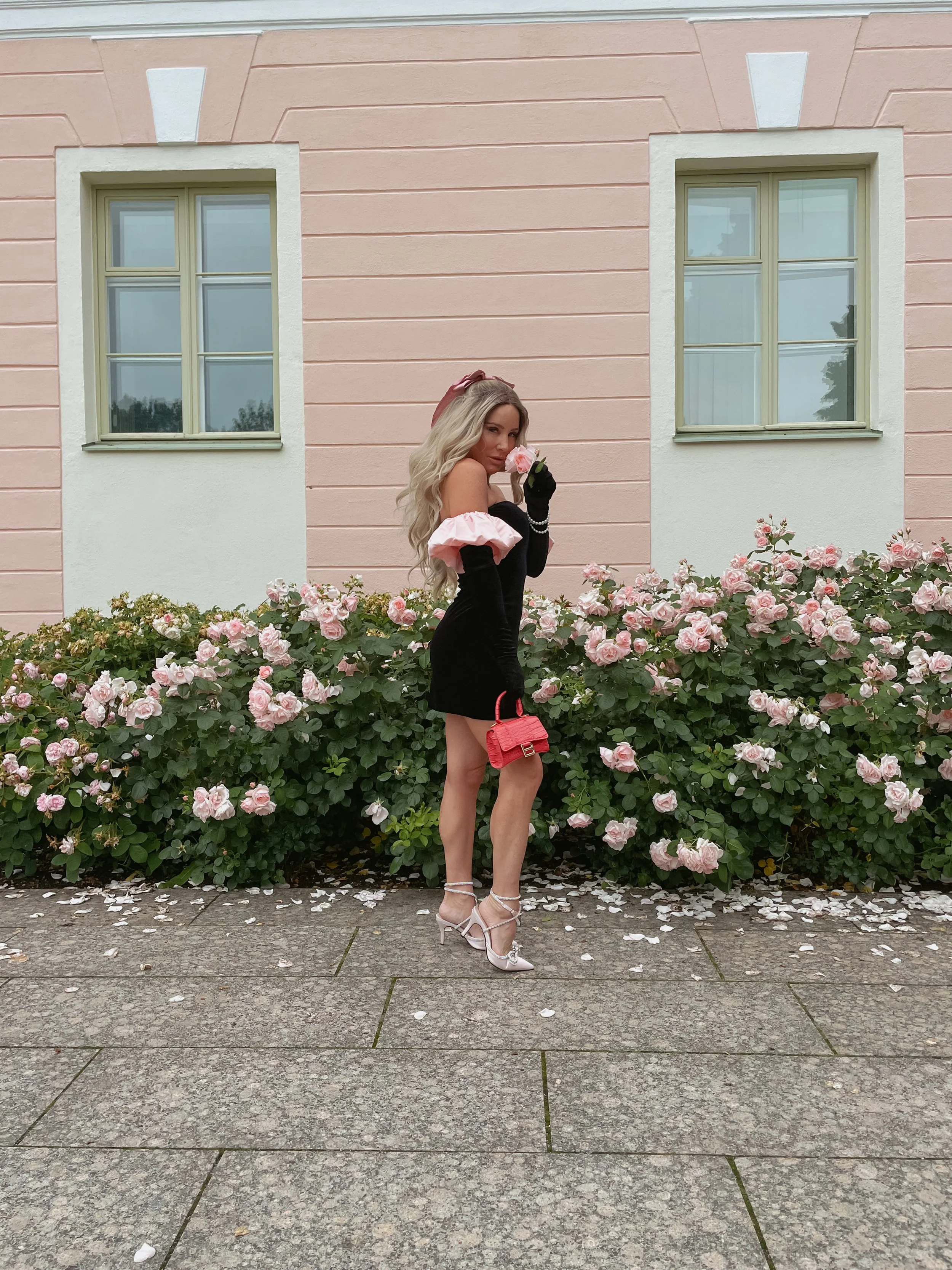 A woman with long blonde hair wearing a black off-shoulder dress with pink ruffles, black gloves, and white high heels, holding a pink rose and standing in front of a building with pink and white walls, surrounded by pink roses.