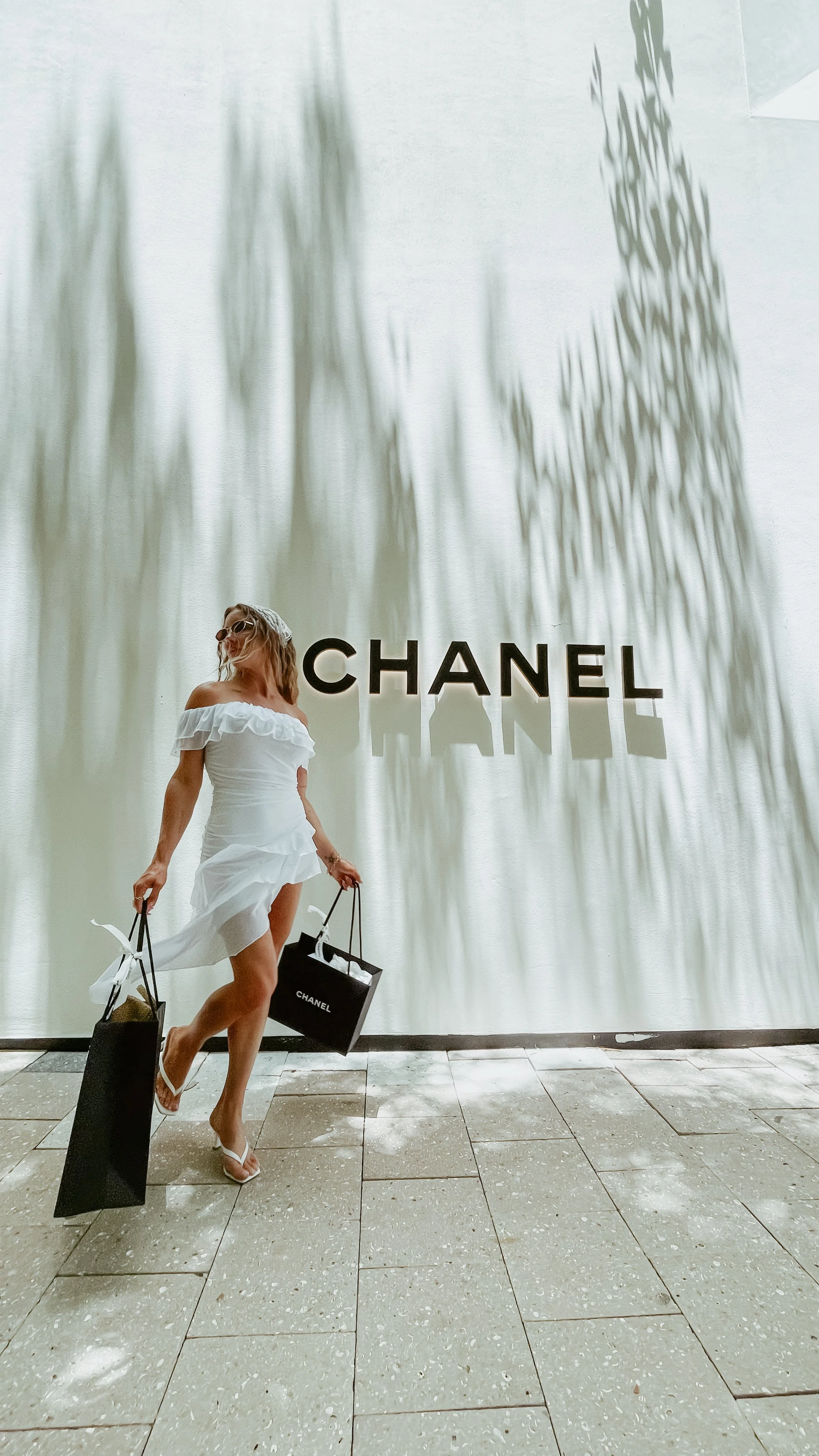 A woman in a white dress with ruffles, sunglasses, and white sandals, holding multiple shopping bags, walking in front of a Chanel store display with large black logo letters and shadows of tree branches on the wall.