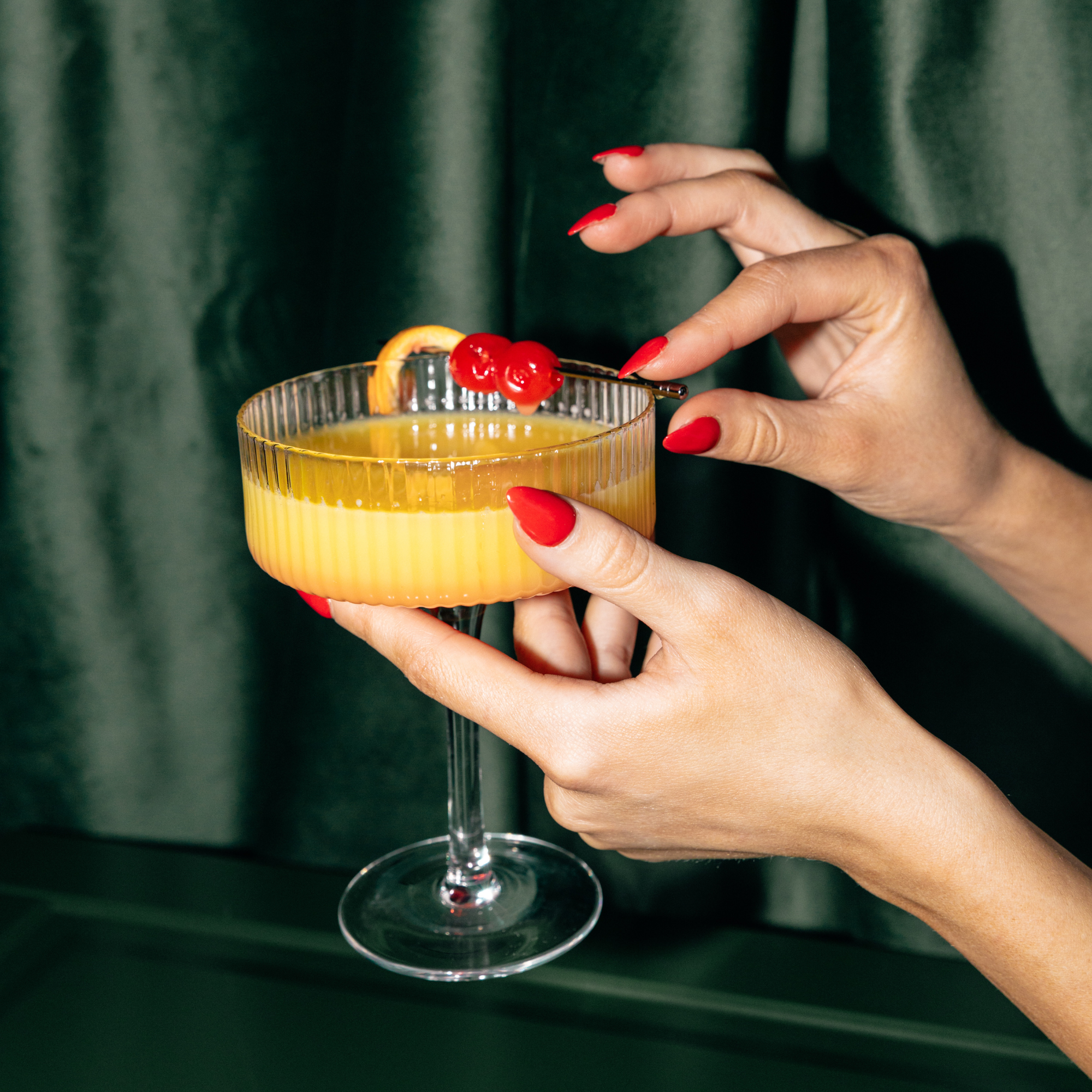 A person with red painted nails holding a cocktail glass with a yellow-orange drink, garnished with cherries and an orange slice, against a dark green background.
