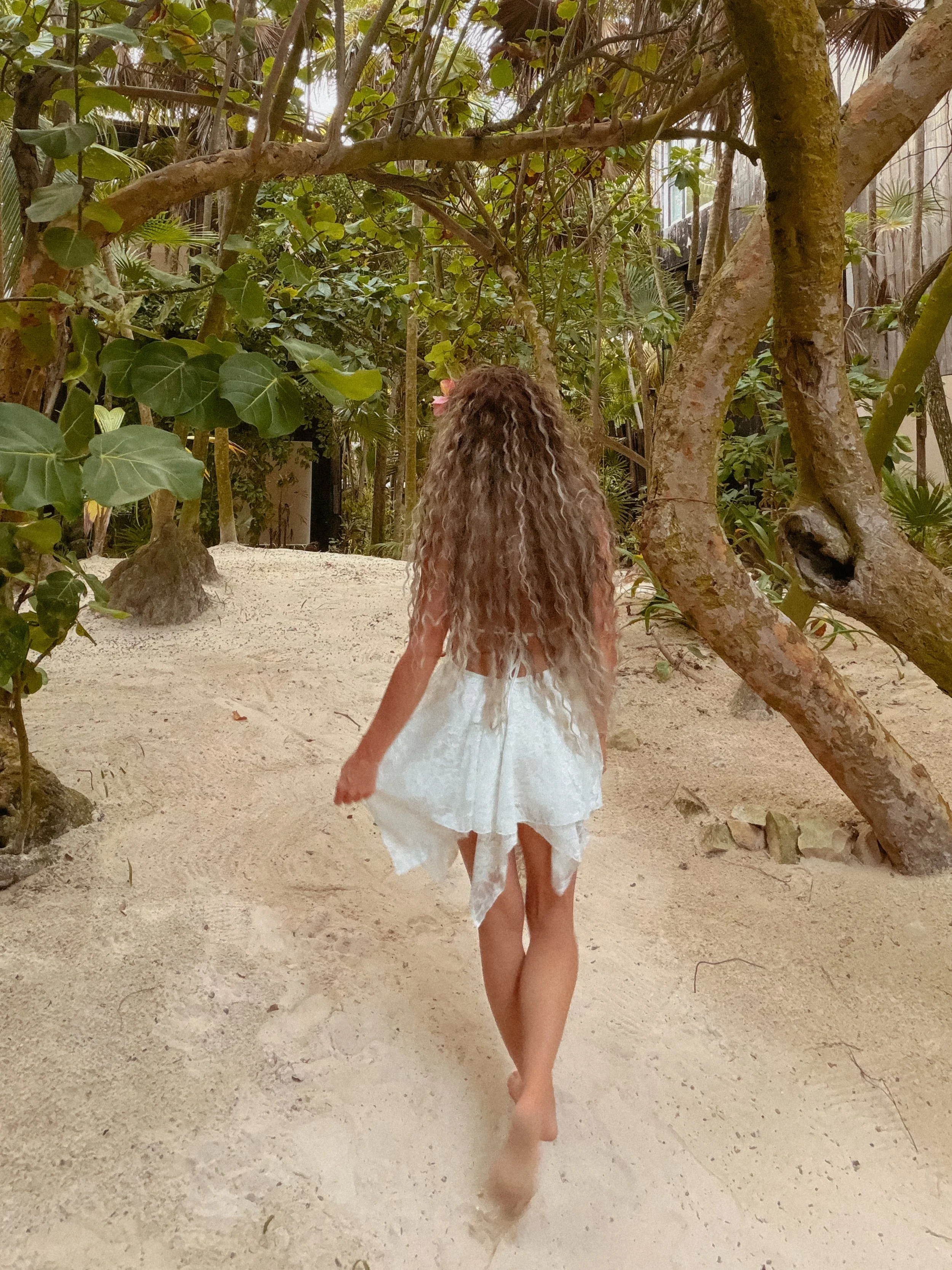 A young girl with curly blonde hair walking barefoot on sandy ground in a lush, green jungle or garden, dressed in a white dress.
