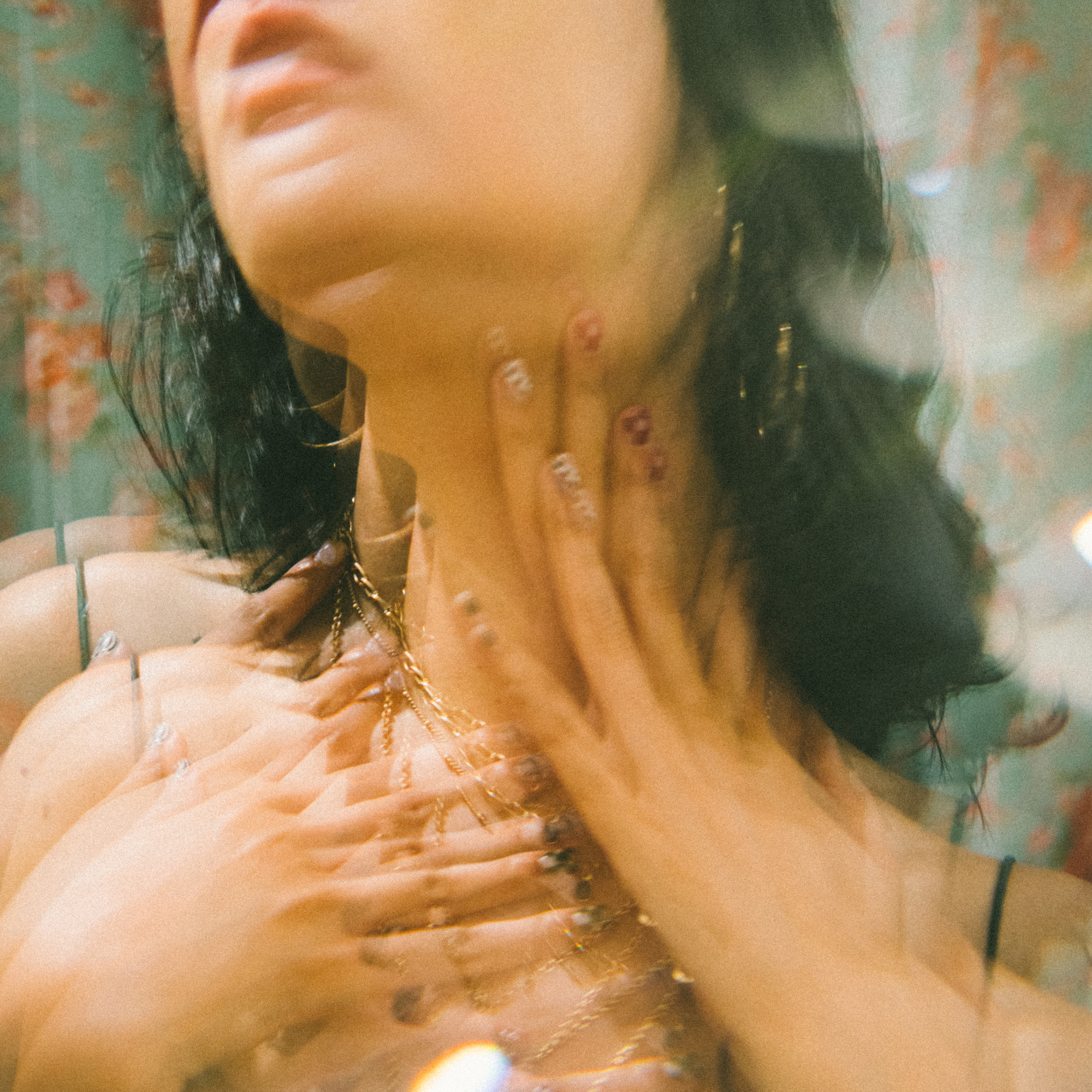 Close-up of a woman's face and hand with rings, gold jewelry, dark hair, and a blurred colorful background.