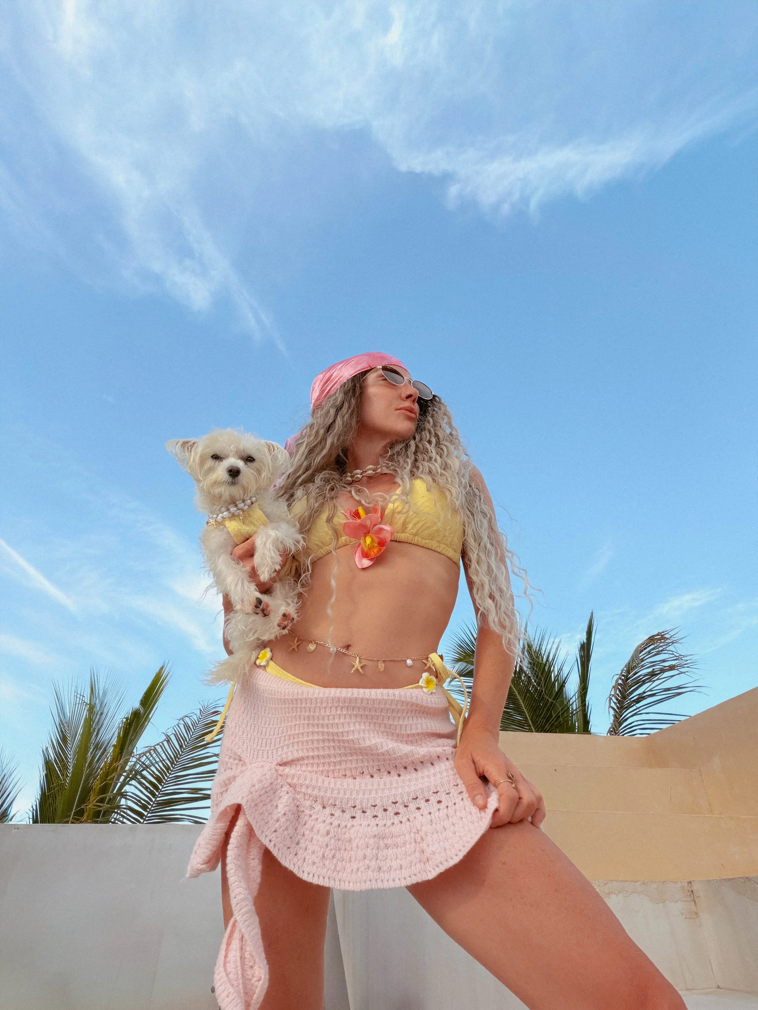 A woman with curly hair wearing sunglasses, a pink headscarf, and a yellow bikini top, holding a small white dog, standing outdoors with palm trees and a blue sky with clouds in the background.