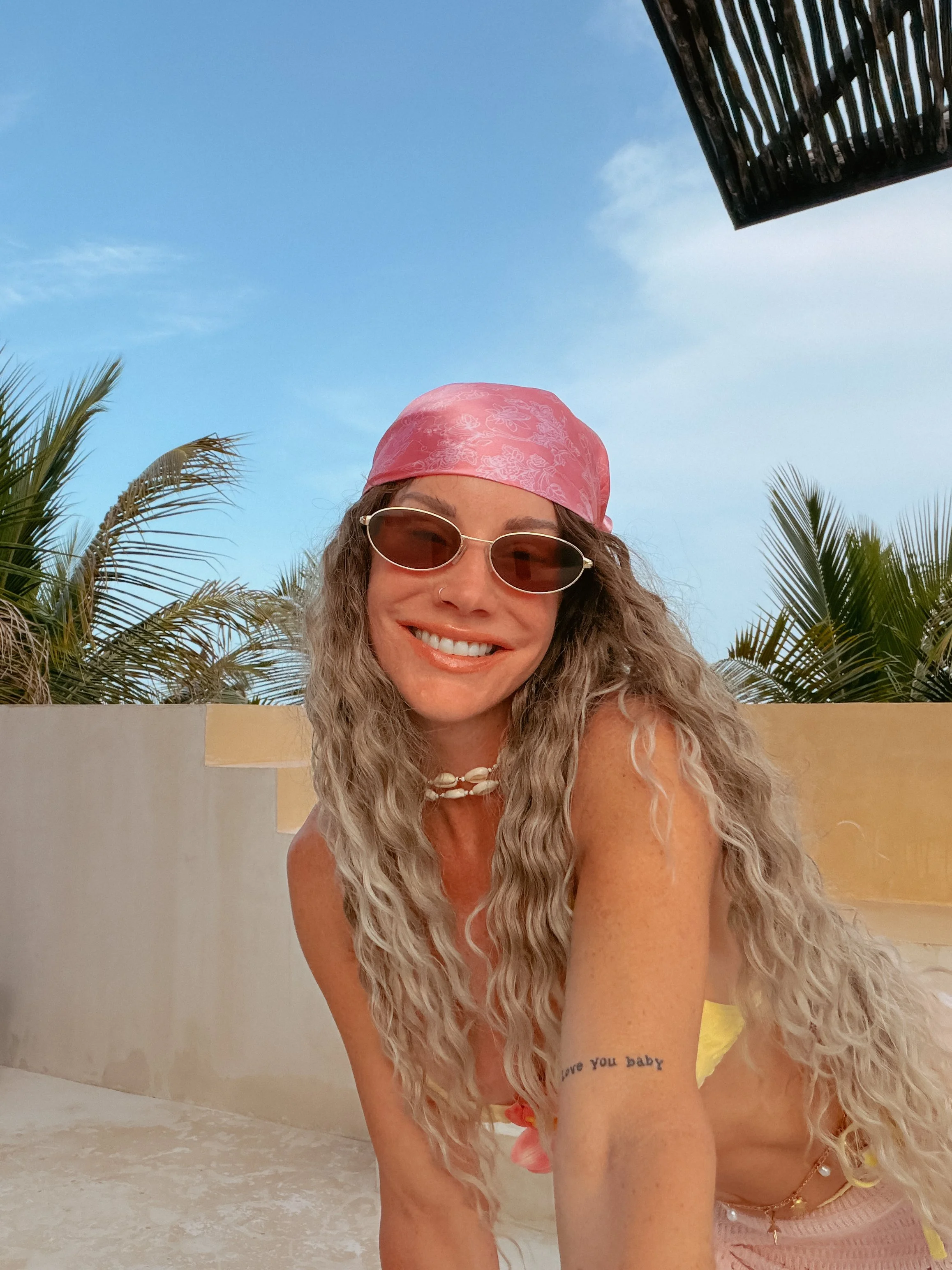A woman with long, wavy blonde hair wearing sunglasses, a pink headscarf, and jewelry, smiling outdoors against a backdrop of palm trees and a blue sky.