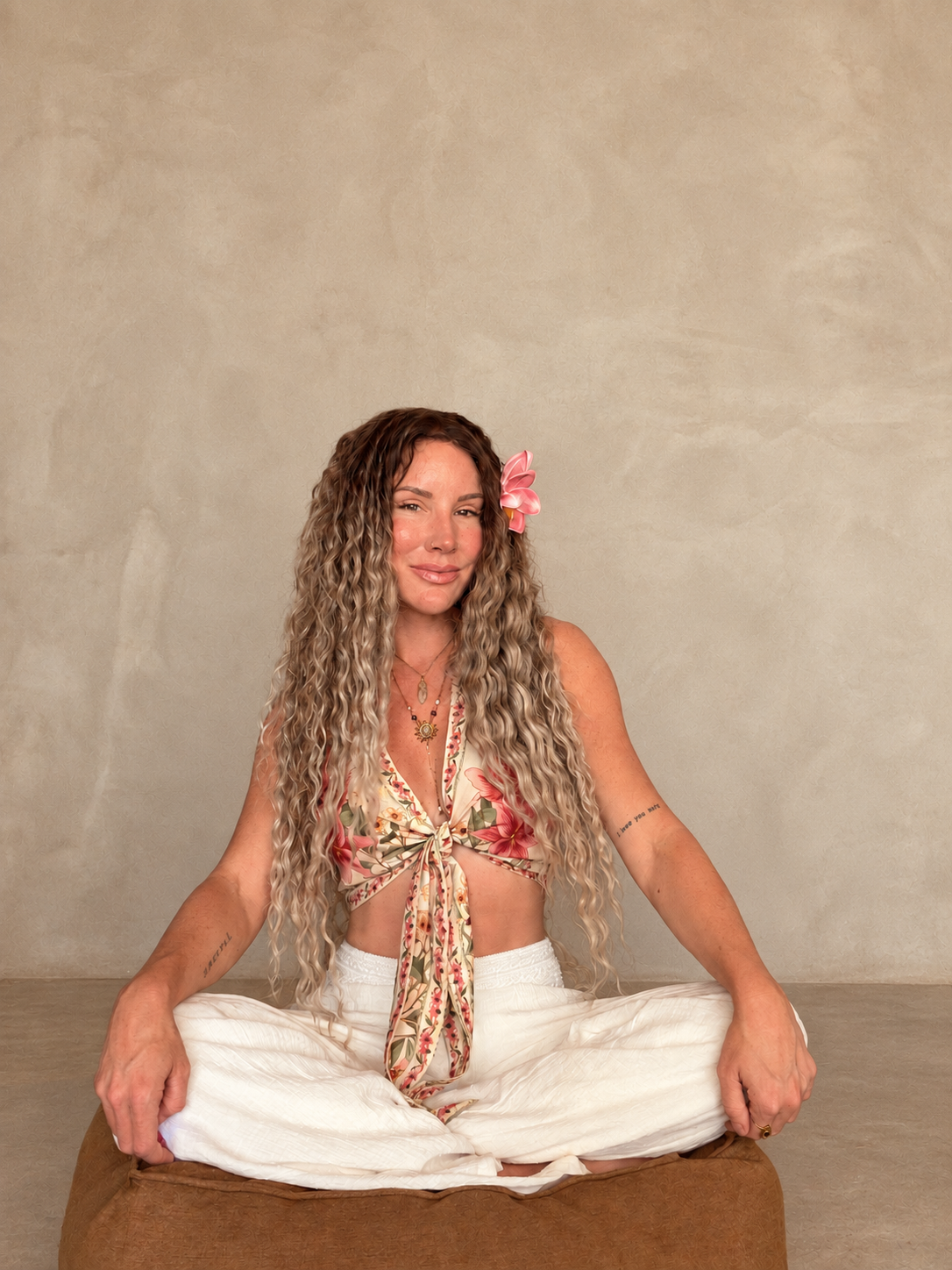 A woman with long curly hair, wearing a floral top with a bow, white pants, and a pink flower in her hair, sitting cross-legged on a cushion in front of a plain beige wall.