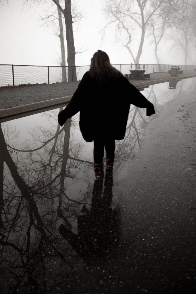 Person walking on a wet, reflective sidewalk during foggy weather in a park with trees and benches in the background.