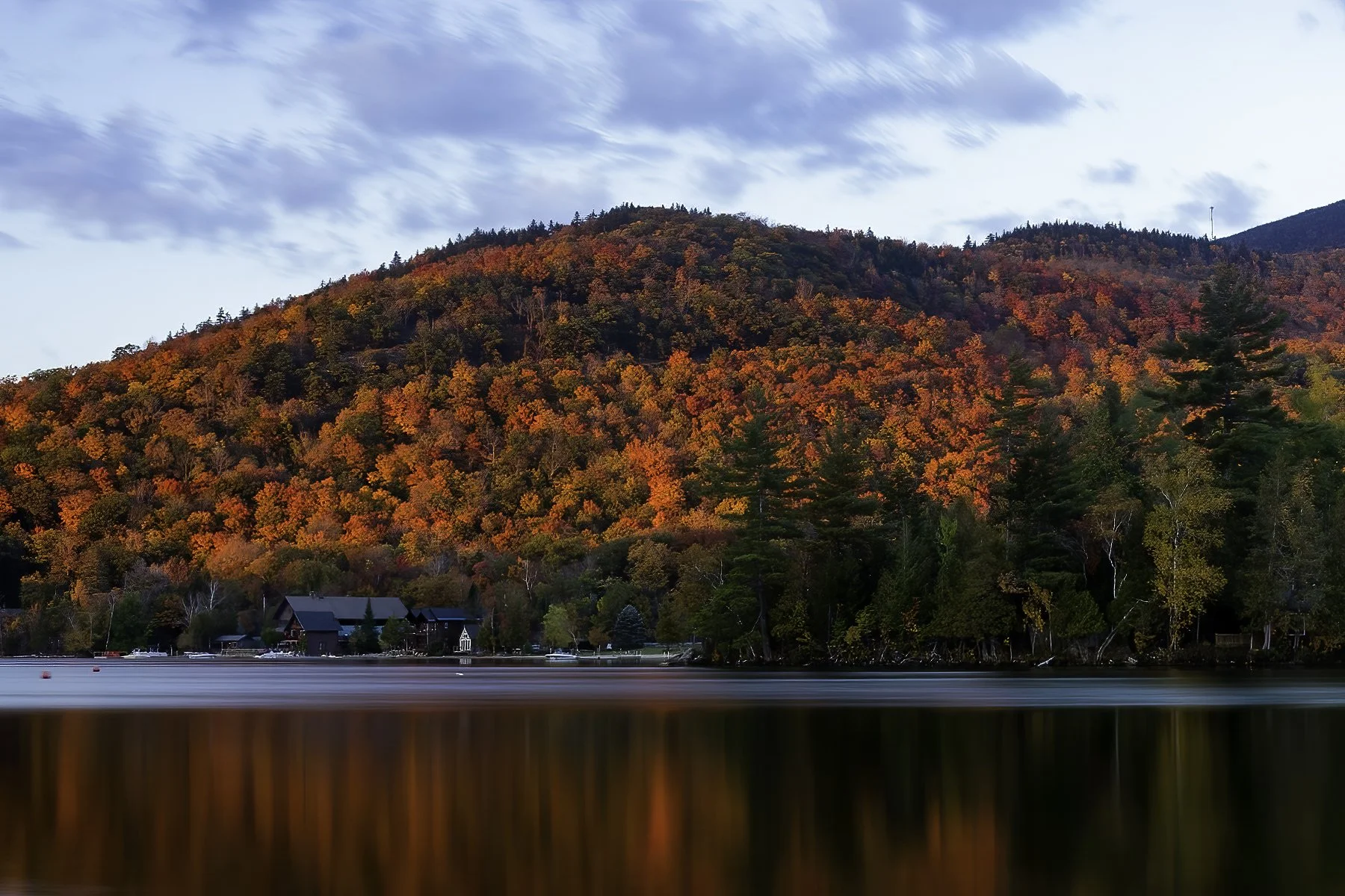 Colorful autumn trees covering a hillside with a few houses near the water's edge in the foreground.
