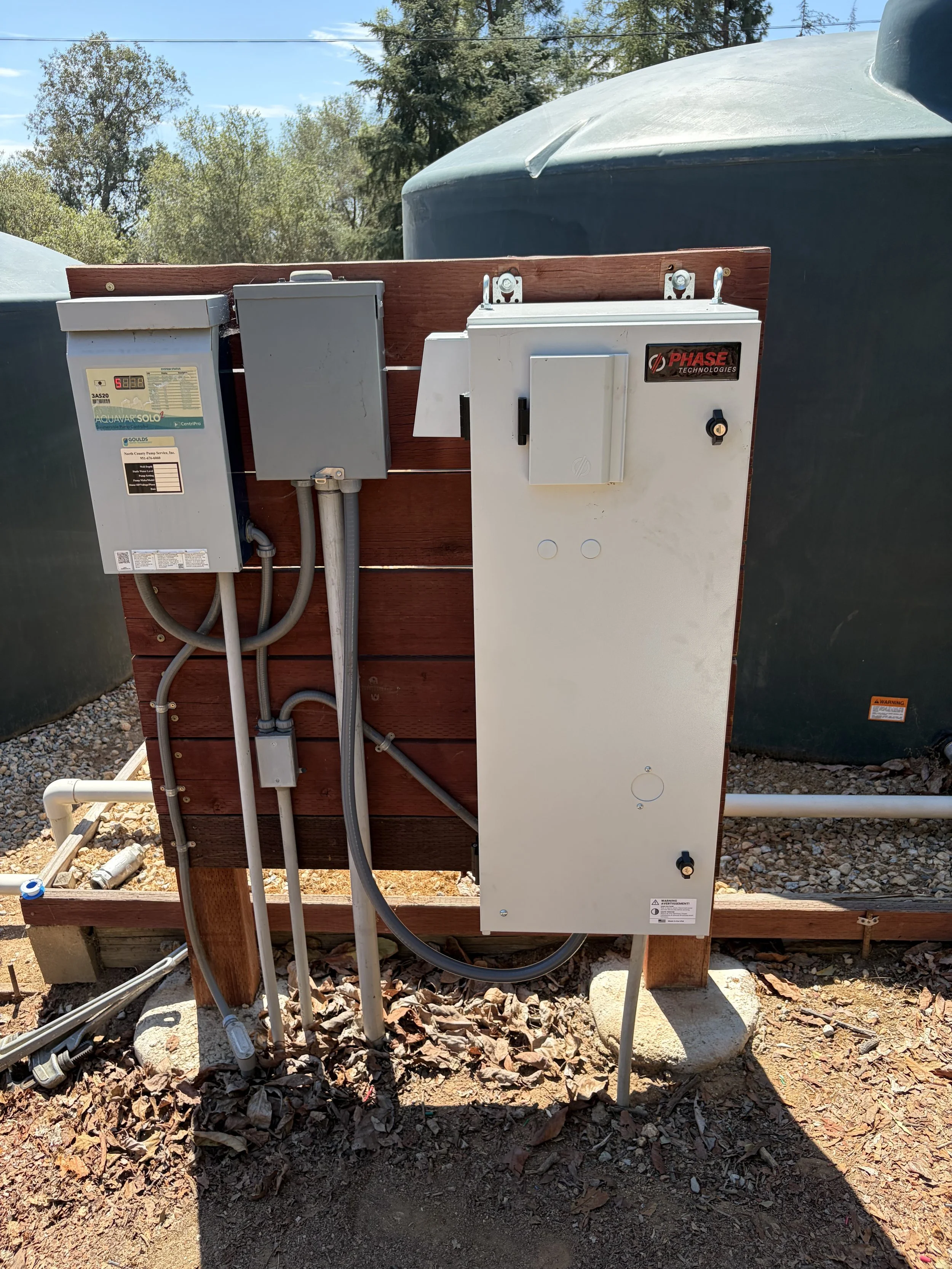 Electrical and control units mounted on a wooden board outside, with water tanks in the background, on gravel ground, in daylight.