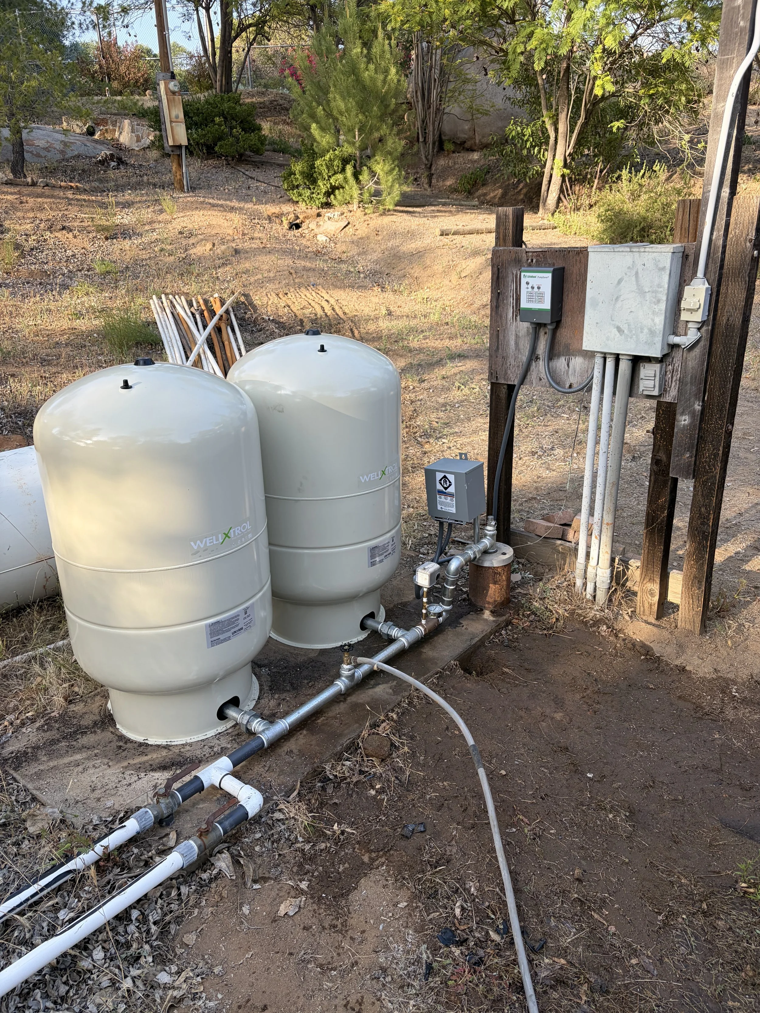 Outdoor scene showing two large white Well-X-Trol water pressure tanks connected to plumbing pipes, with a control panel and electrical components mounted on a wooden post, in a yard with dirt, small plants, trees, and a fence in the background.
