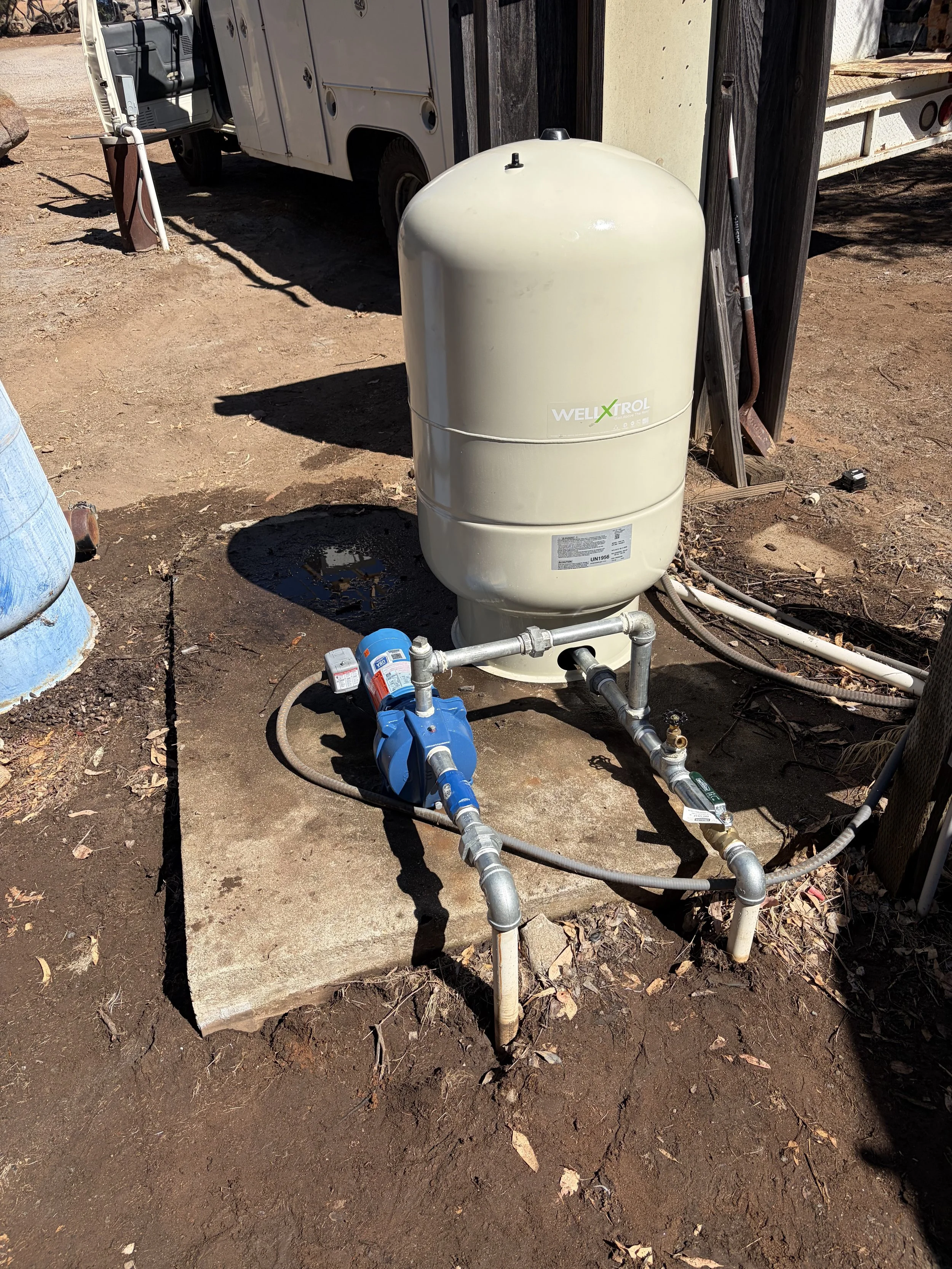 A well system with pipes and a beige well tank installed on dirt ground, next to a wooden fence and a parked white utility vehicle.