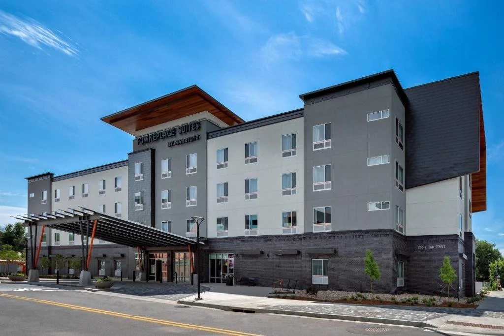 A modern four-story apartment building named 'Townplace Suites by Marriott' with a large slanted roof extension, glass entrance, and small trees along the sidewalk, under a blue sky.
