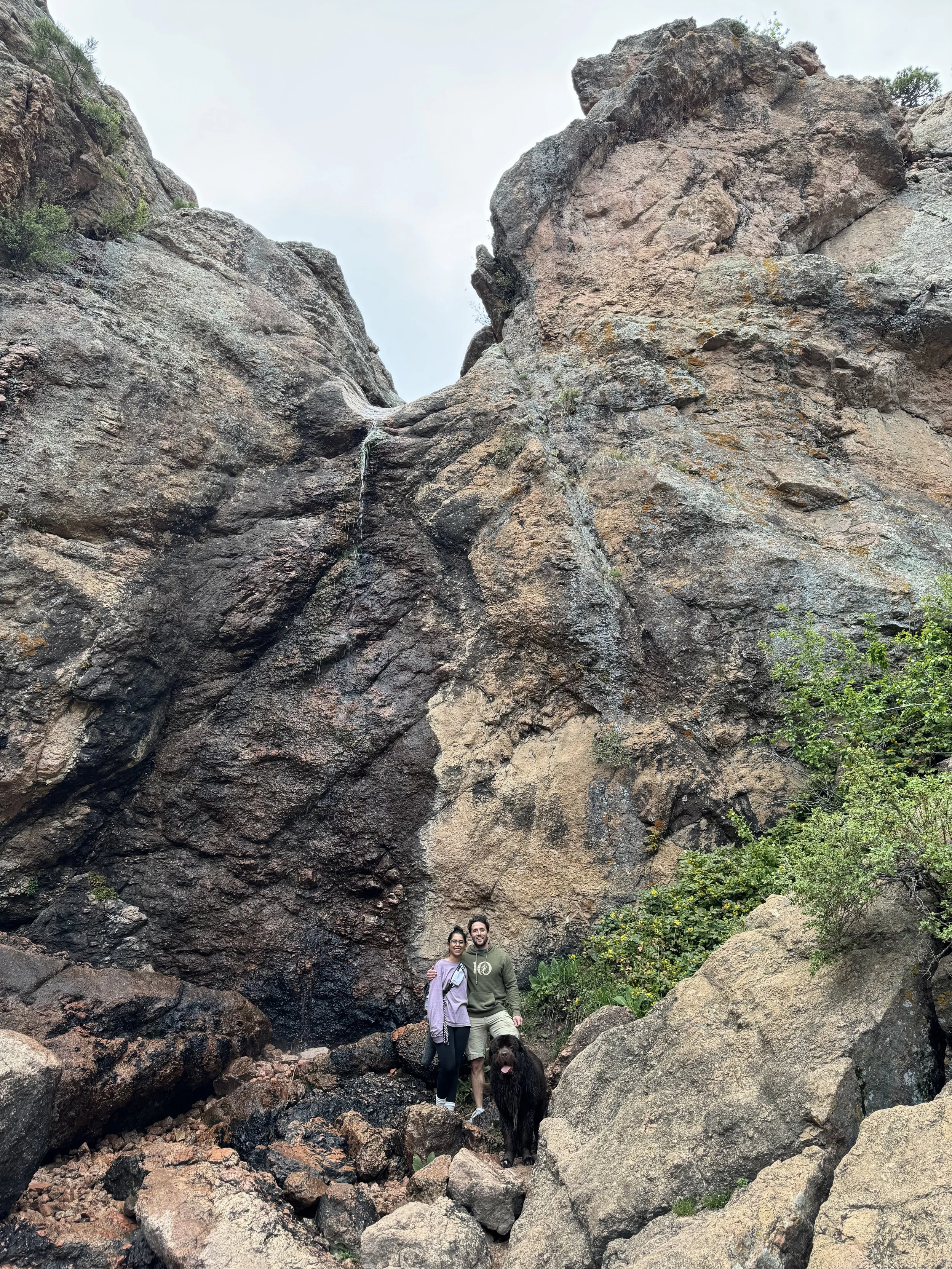 Two people and a dog standing on rocky terrain in front of a large rock formation, with greenery around.