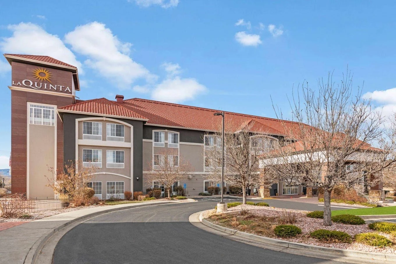 Exterior view of a La Quinta hotel with a curved driveway, leafless trees, and a bright blue sky with clouds.