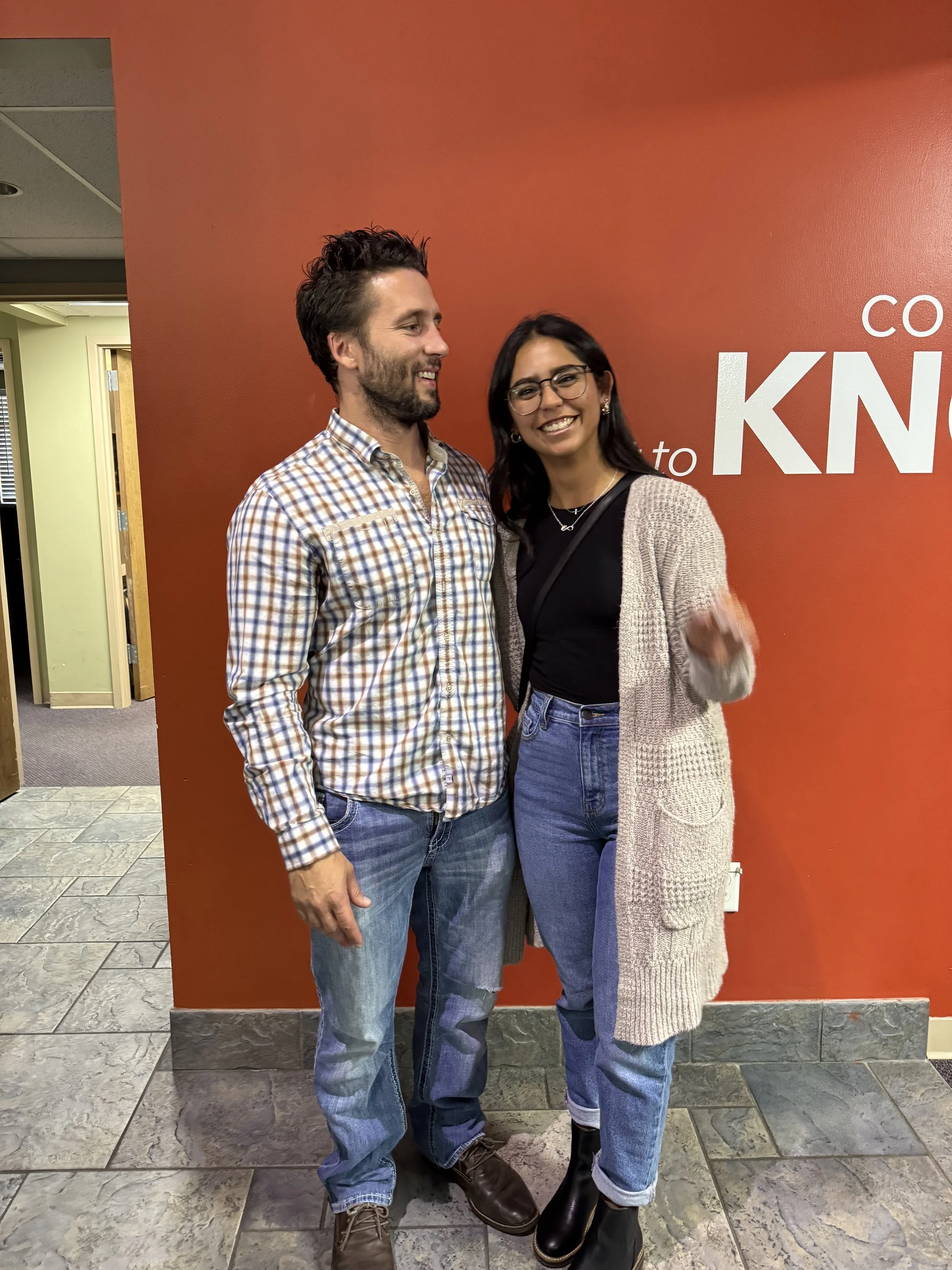 A man and woman standing together smiling in front of a red wall with white text. The man is wearing a plaid shirt and jeans, the woman is wearing glasses, a black top, jeans, and a beige cardigan.