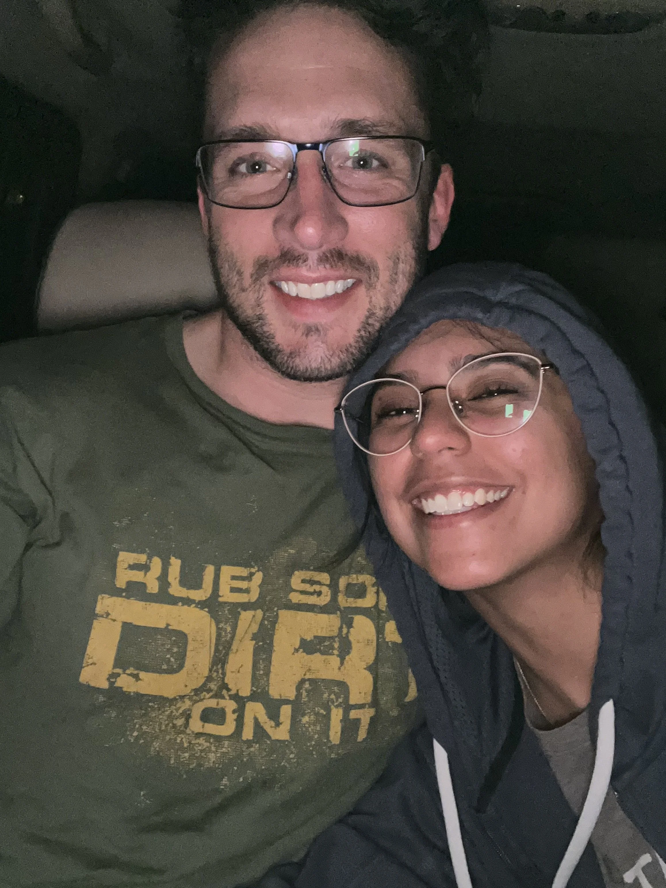 Close-up of a smiling man with glasses, a beard, and short hair, wearing a green t-shirt. Part of a woman with glasses and a hooded jacket is visible, along with a shirt that says 'FUB' and 'ON T' in yellow. The background appears to be the interior 