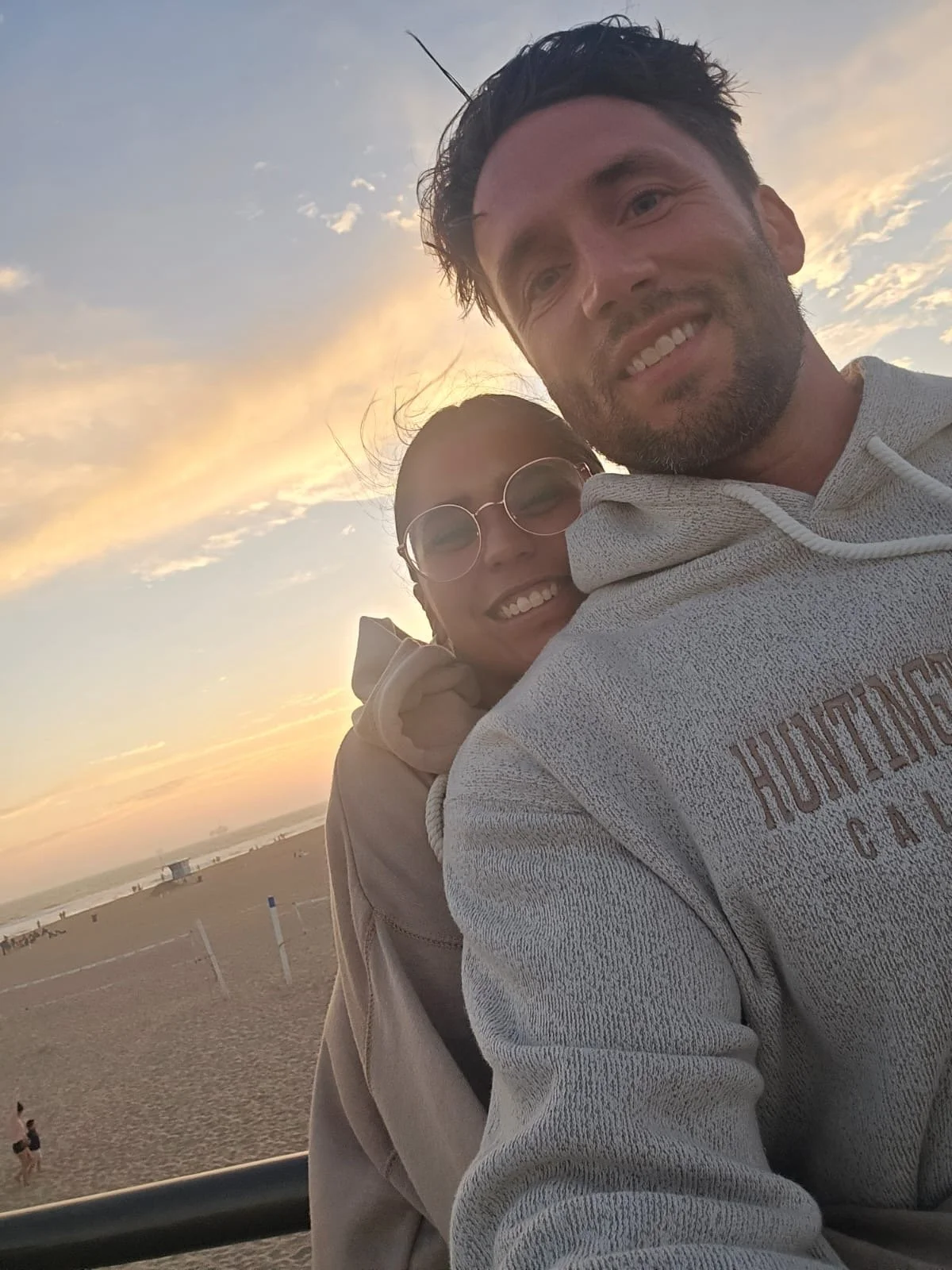A man and woman taking a selfie on the beach at sunset, with the ocean and a few people in the background.