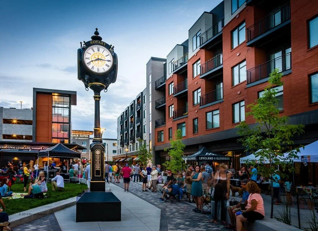 A busy outdoor urban scene at dusk with a large vintage-style clock on a tall black pole, surrounded by modern apartment buildings and street vendors, people walking, sitting, and socializing.