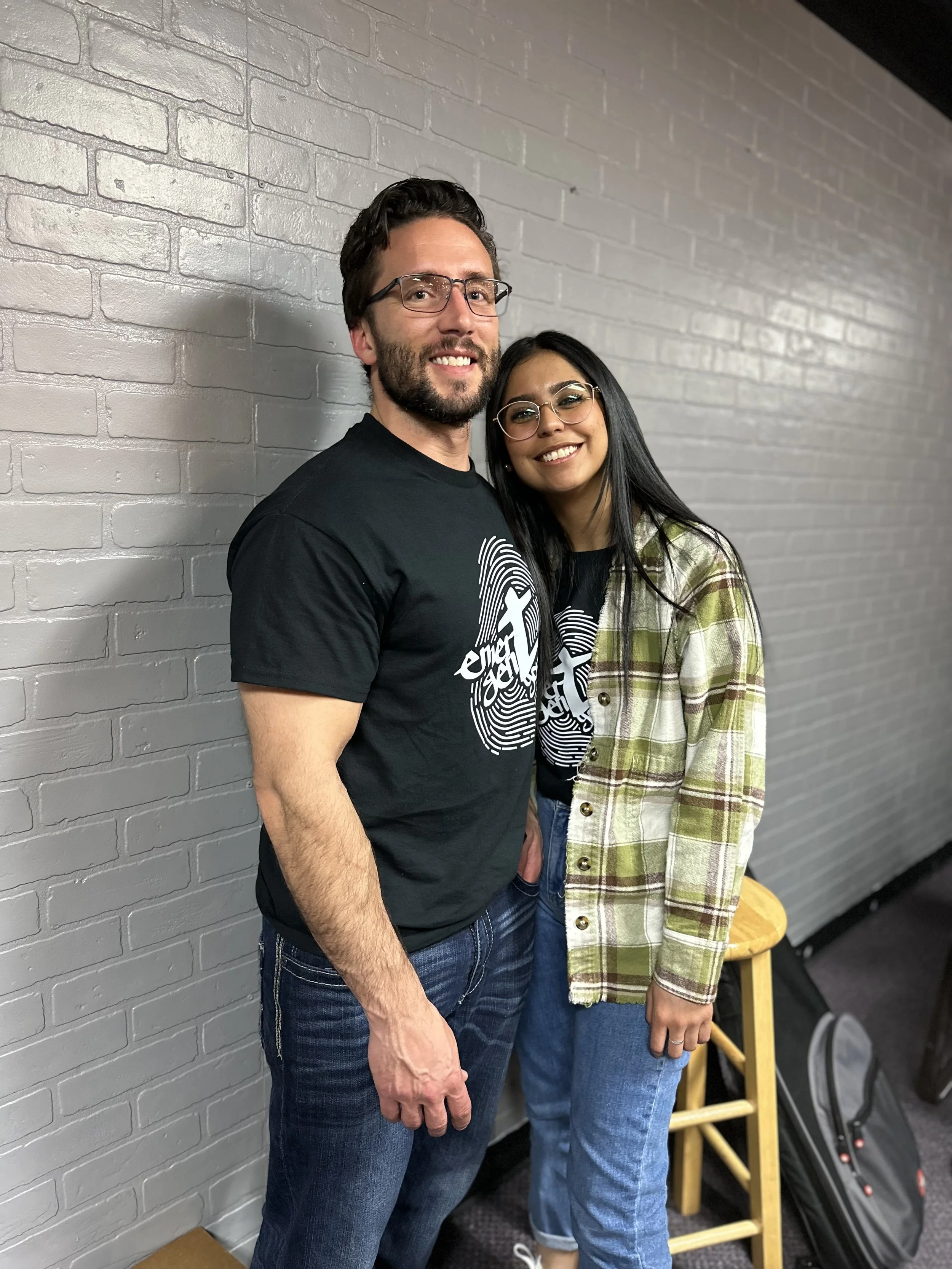 A man and a woman are standing close together, smiling and posing for a photo against a gray brick wall. The man has dark hair, beard, and glasses, wearing a black t-shirt. The woman has long dark hair, glasses, and is wearing a plaid shirt over a black t-shirt and jeans.