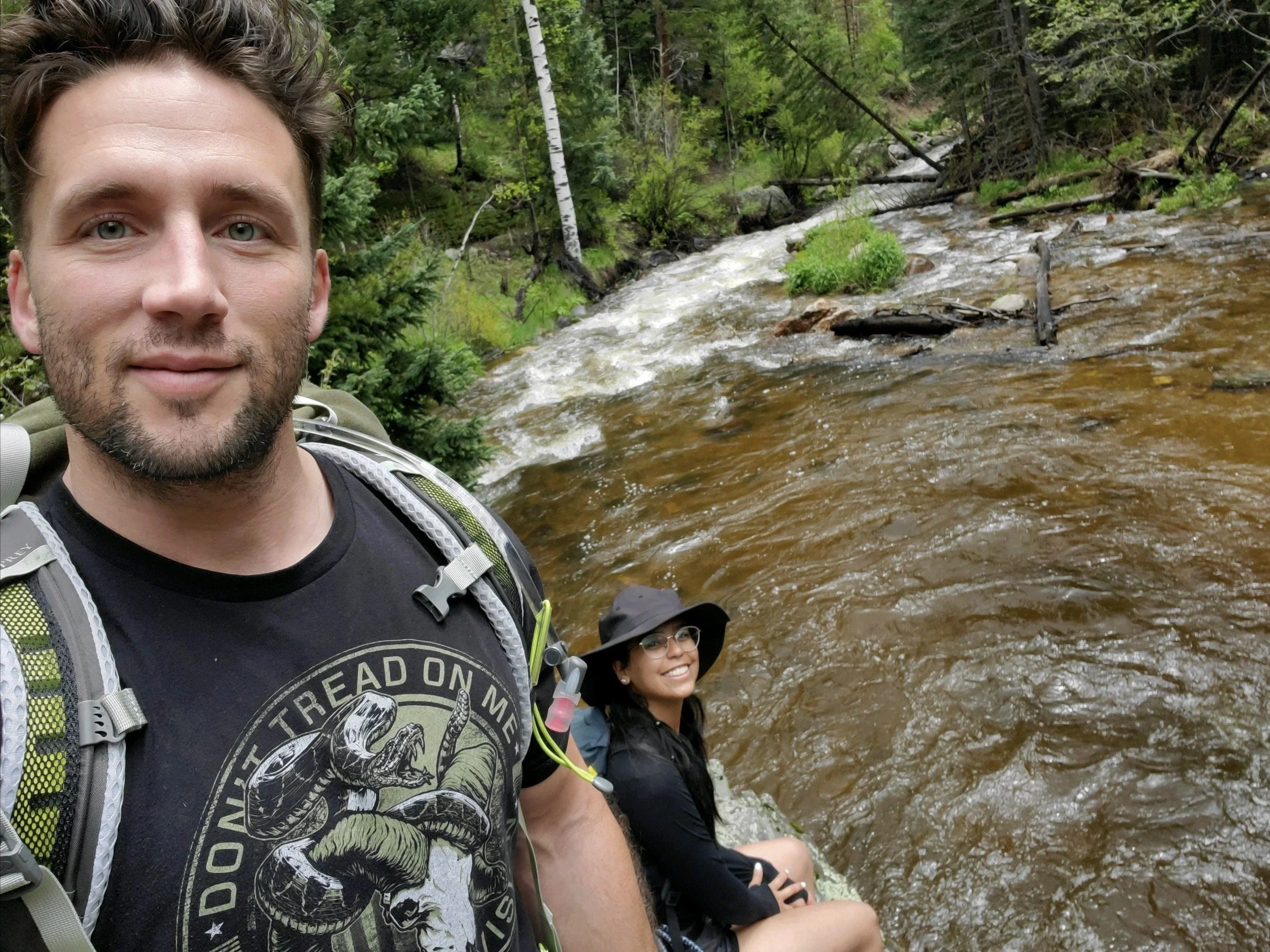 A man and a woman hiking by a stream in a lush forest.