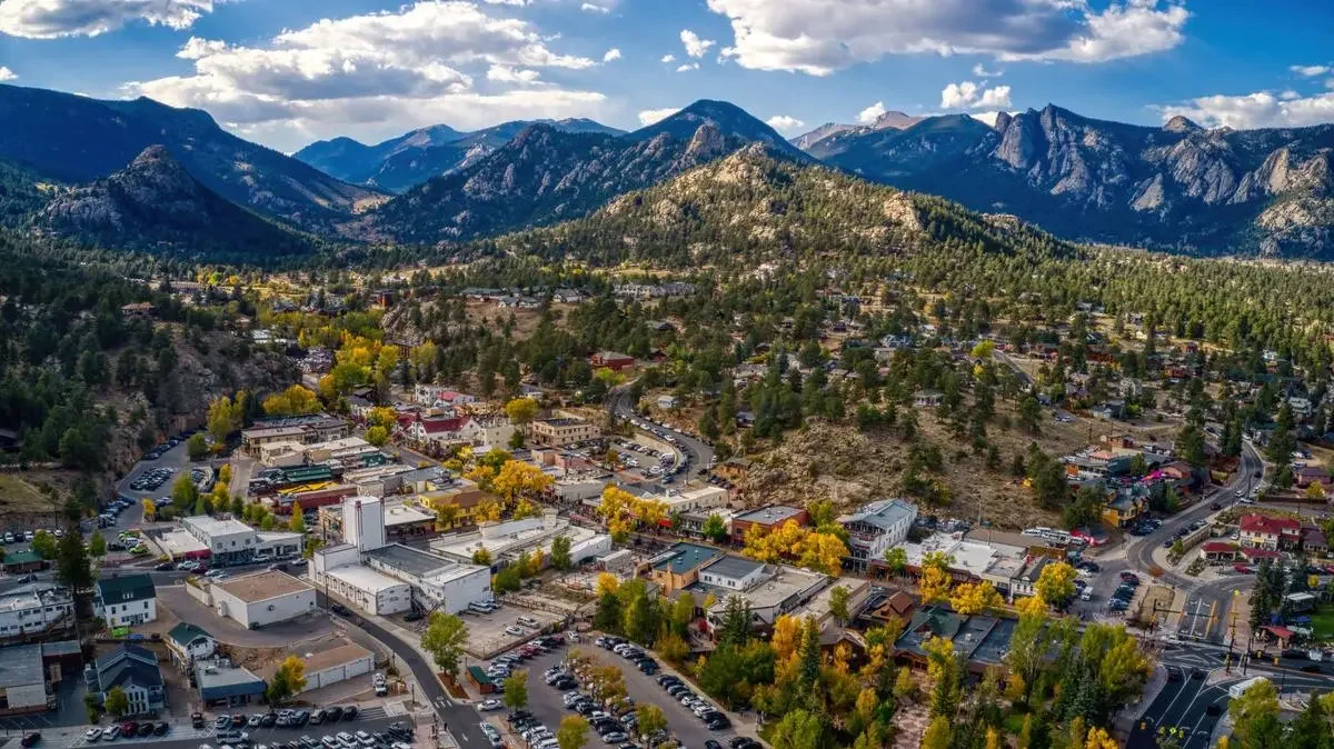 Aerial view of a mountain town with colorful trees, buildings, and parking lots, surrounded by forested hills and mountains under a partly cloudy sky.
