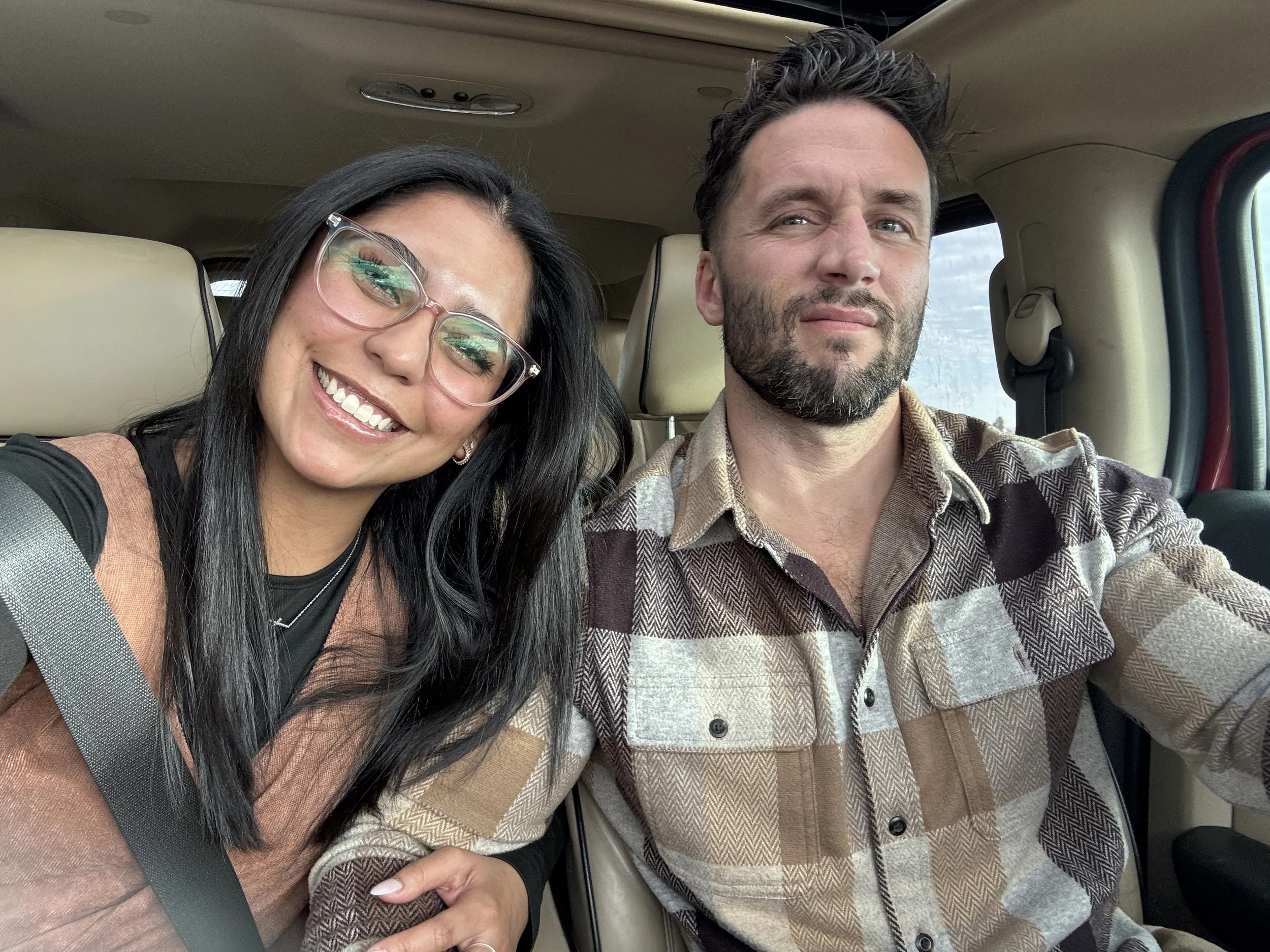 A woman and a man sitting in a vehicle, close together, smiling at the camera. The woman has long black hair, glasses, and is wearing a tan and black outfit. The man has dark hair, a beard, and is wearing a plaid shirt.
