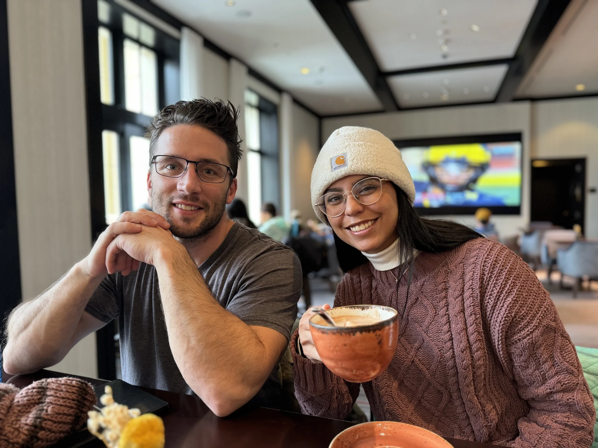 A smiling man with glasses and a woman wearing a beige Carhartt beanie and glasses, sitting together at a table in a restaurant or cafe. They are enjoying drinks, with the woman holding an orange ceramic mug. The background shows a modern interior wi